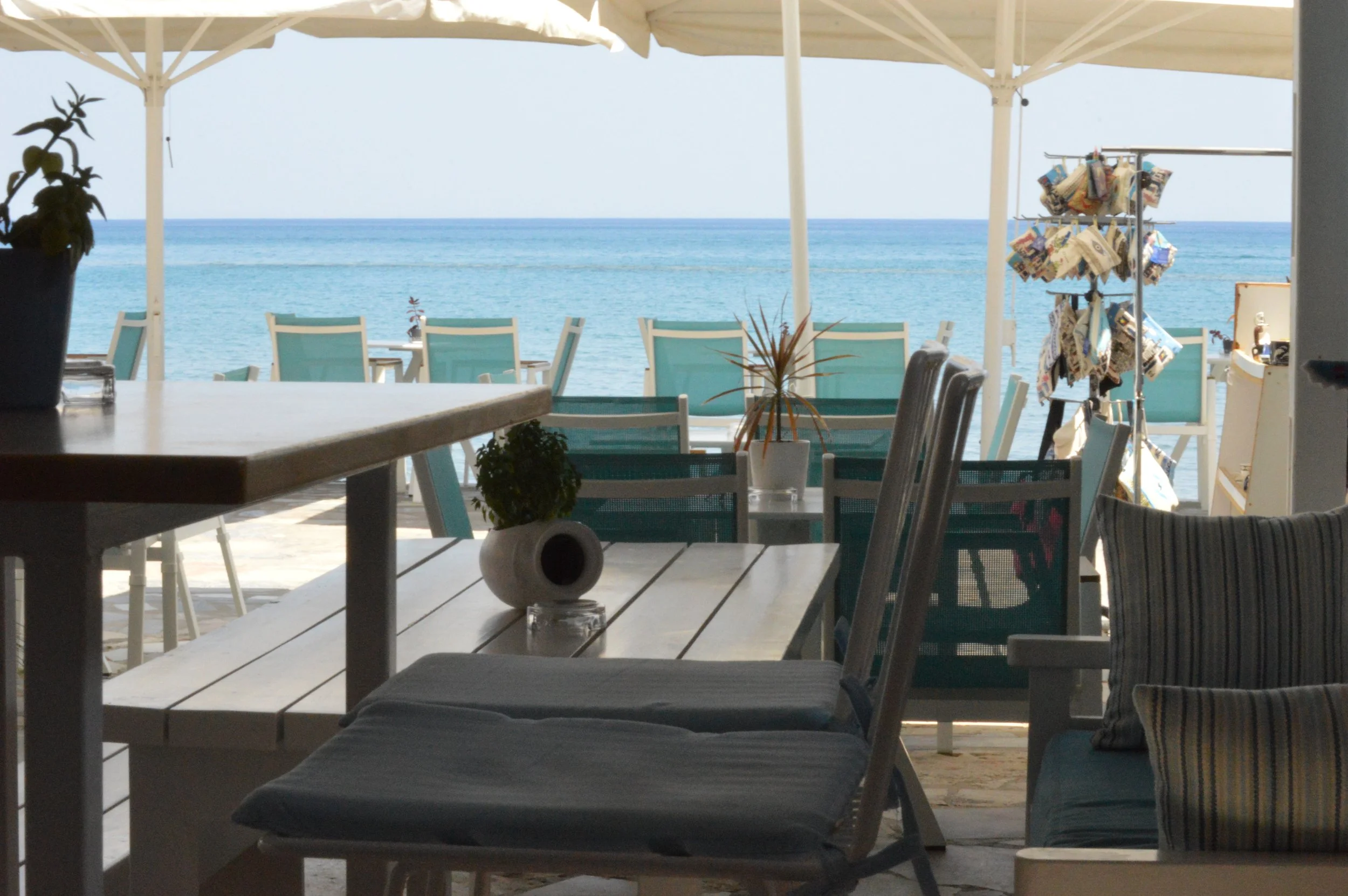Gelateria Maria - View from inside a beachside cafe or restaurant with a table and chairs, overlooking the ocean with a clear sky, some umbrellas, and a display rack of merchandise outside.