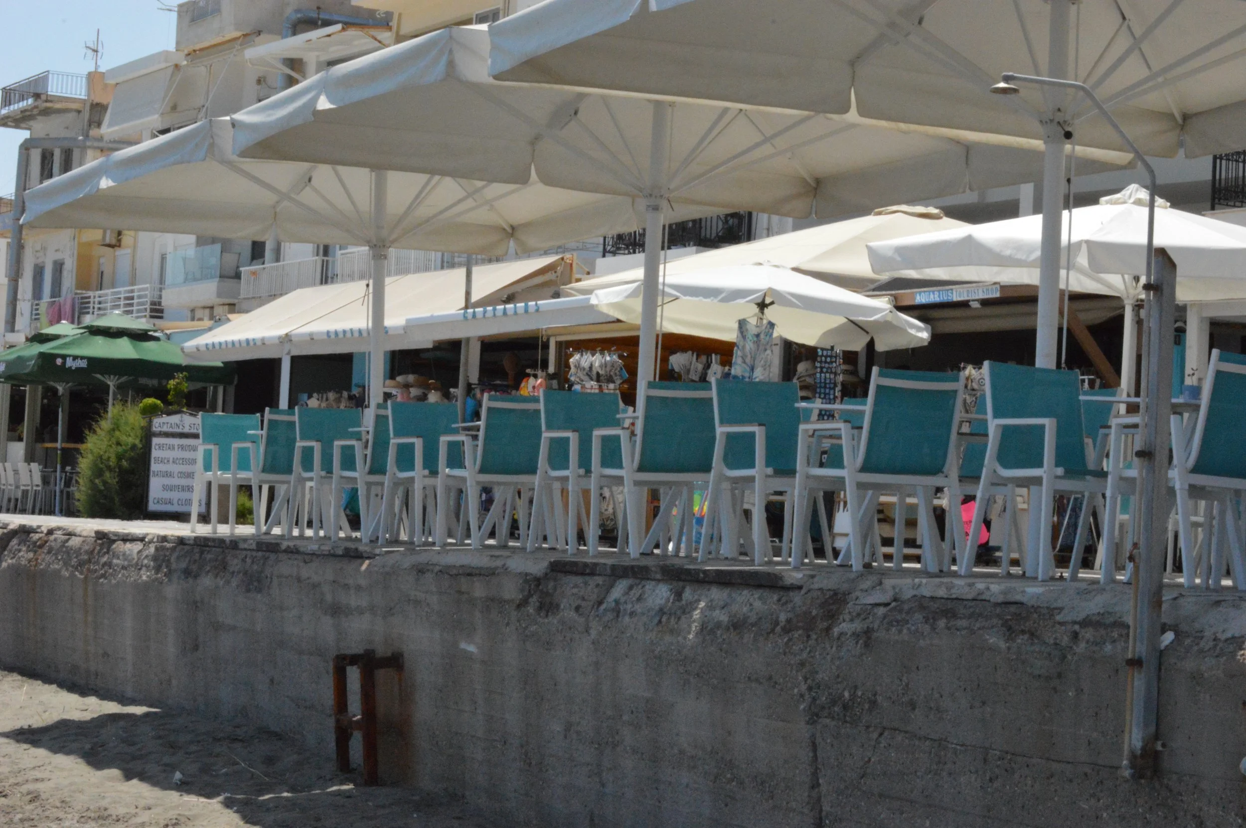 Beachside outdoor seating area with white chairs and umbrellas, near sandy beach and beachfront buildings in the background.