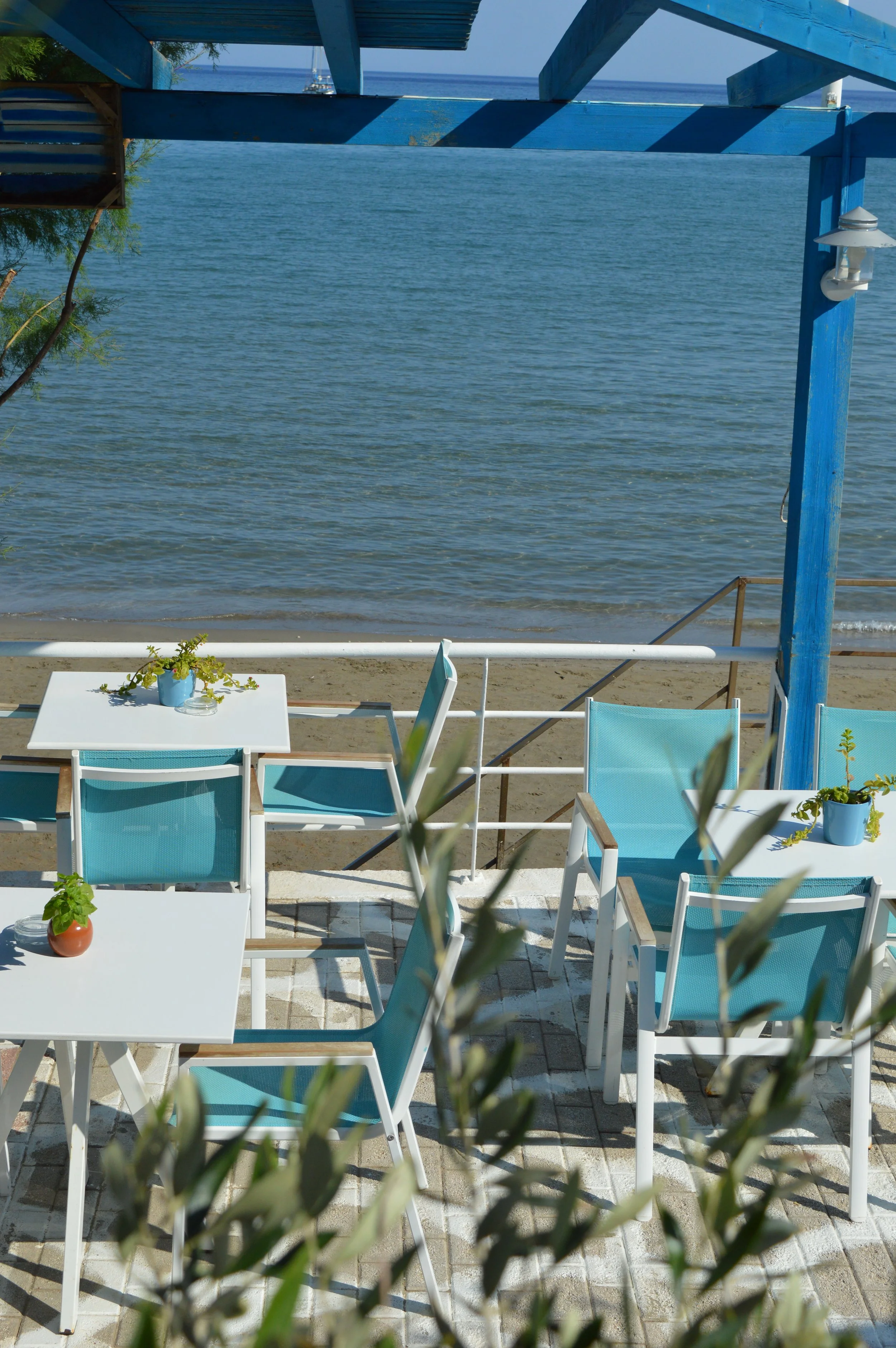 Outdoor patio with white tables and blue chairs overlooking a calm body of water, with a blue-painted structure and some plants in pots.