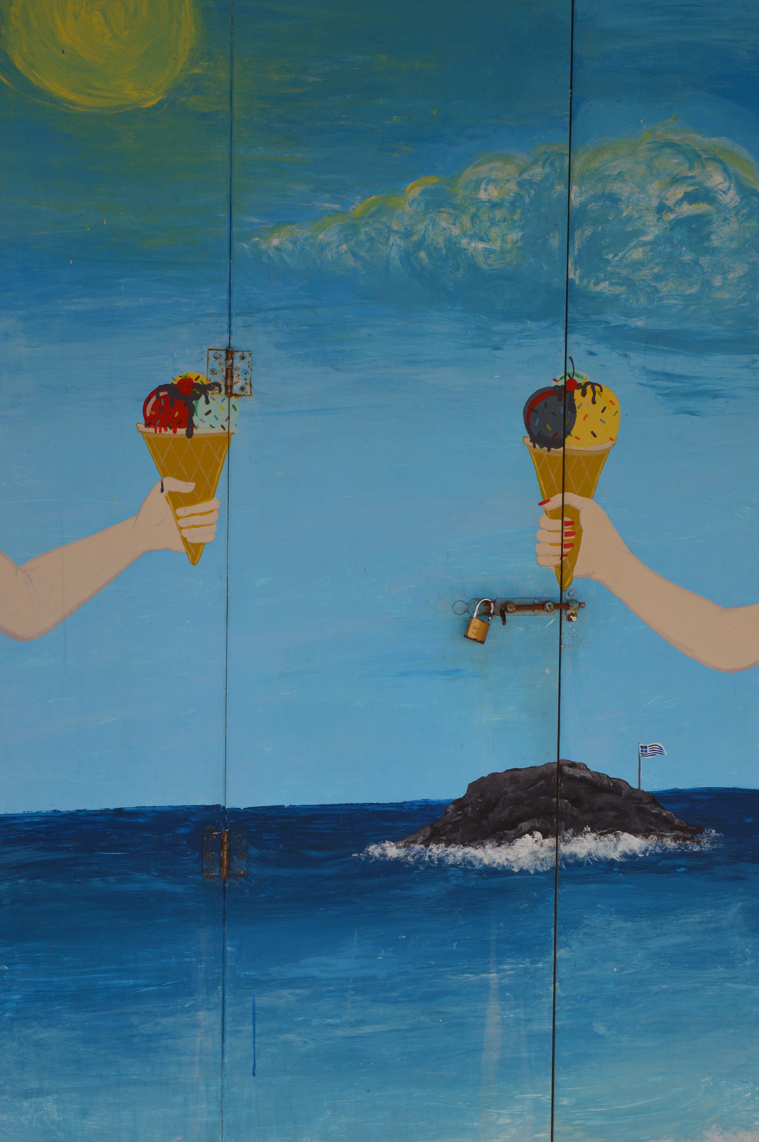 Gelateria Maria - Mural painting of two hands holding ice cream cones in the sky with clouds and a sun, and a small island with a Greek flag in the ocean below.