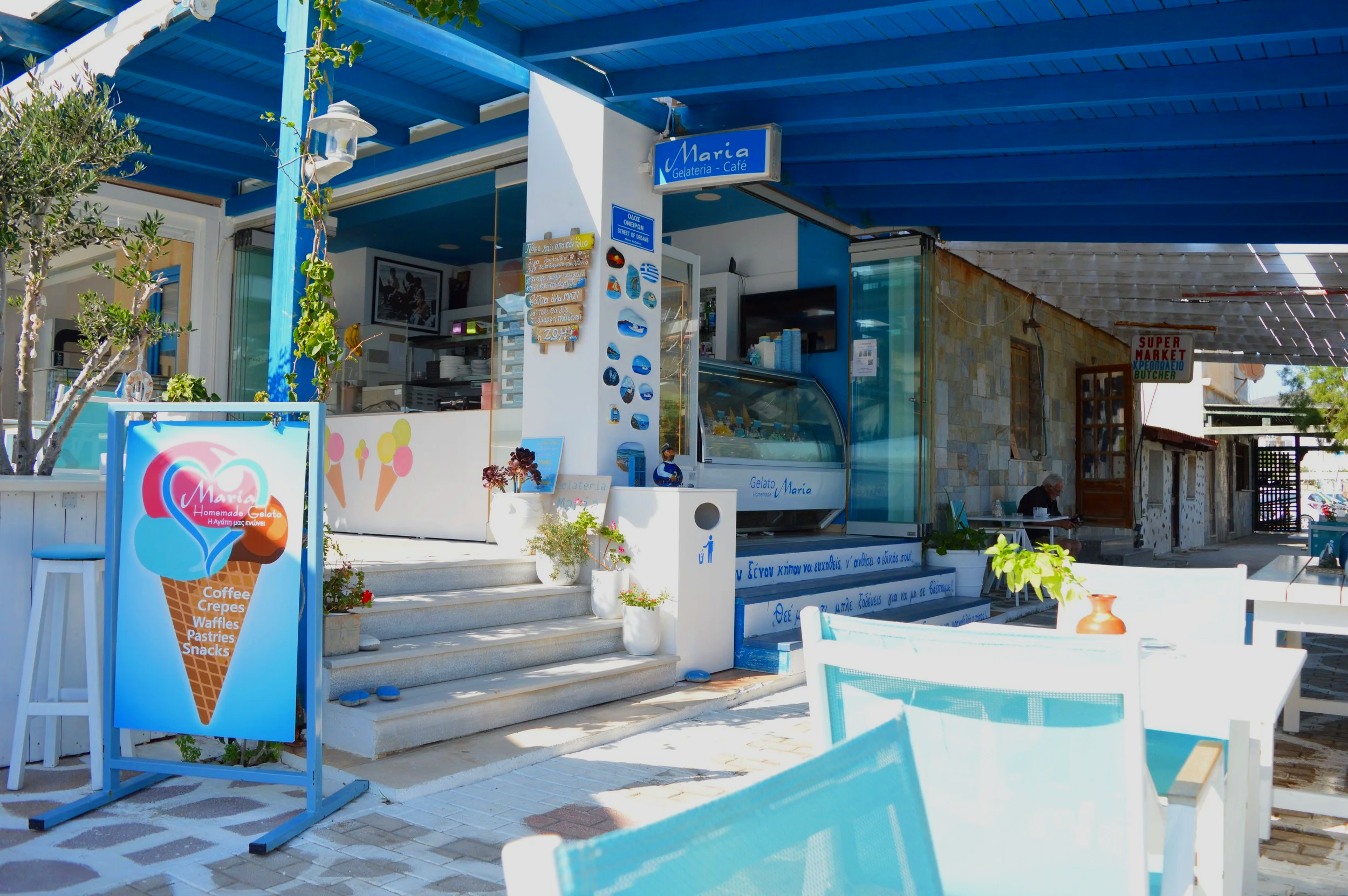 Exterior view of an ice cream shop with a blue awning and sign reading 'Maria Gelateria Café'. There is a colorful ice cream cone signboard listing offerings like coffee, crepes, waffles, pastries, and snacks. The shop has steps leading up to the cou