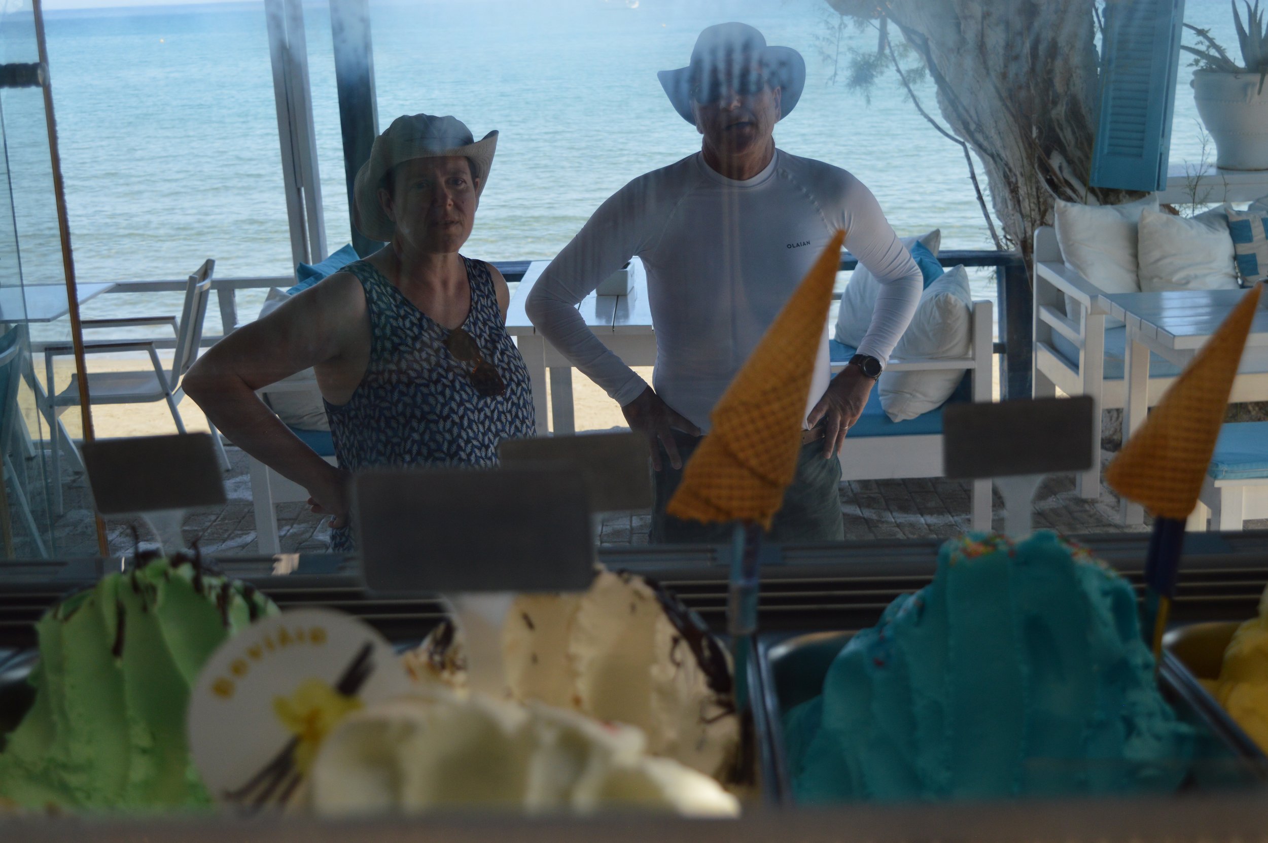 Gelateria Maria - Two people, a woman and a man, looking at ice cream behind a glass window at a beachside location, with water and outdoor seating visible in the background.