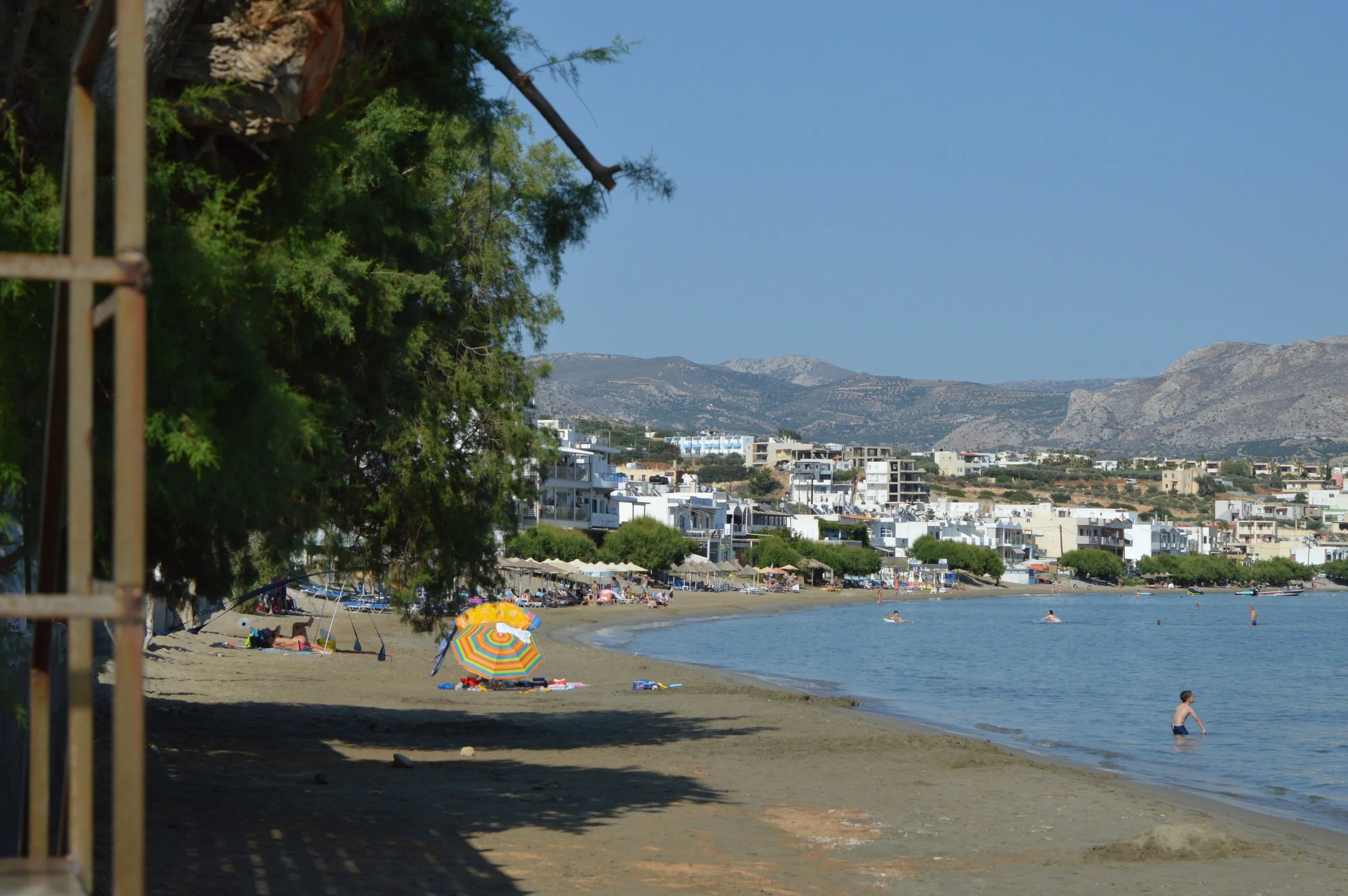 A beach scene with people swimming and relaxing under colorful umbrellas, lined with white buildings and mountains in the background on a sunny day.