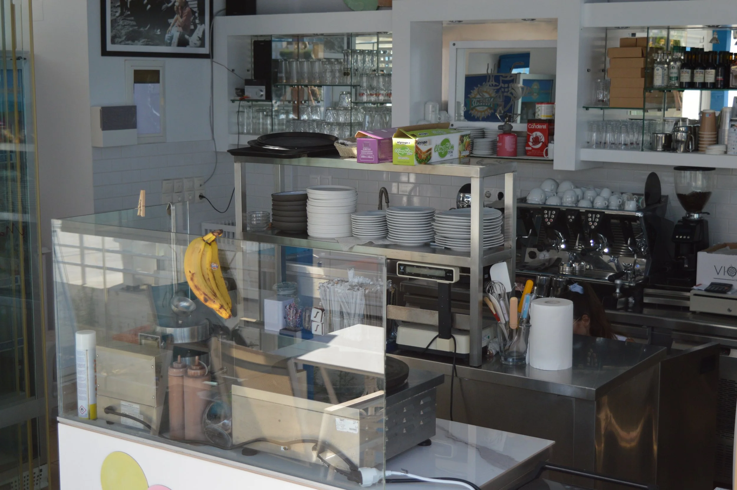 Gelateria Maria - Interior of a coffee shop counter with stacked plates, coffee machine, and various supplies. Bananas hang in the foreground, and glassware is visible on shelves in the background.