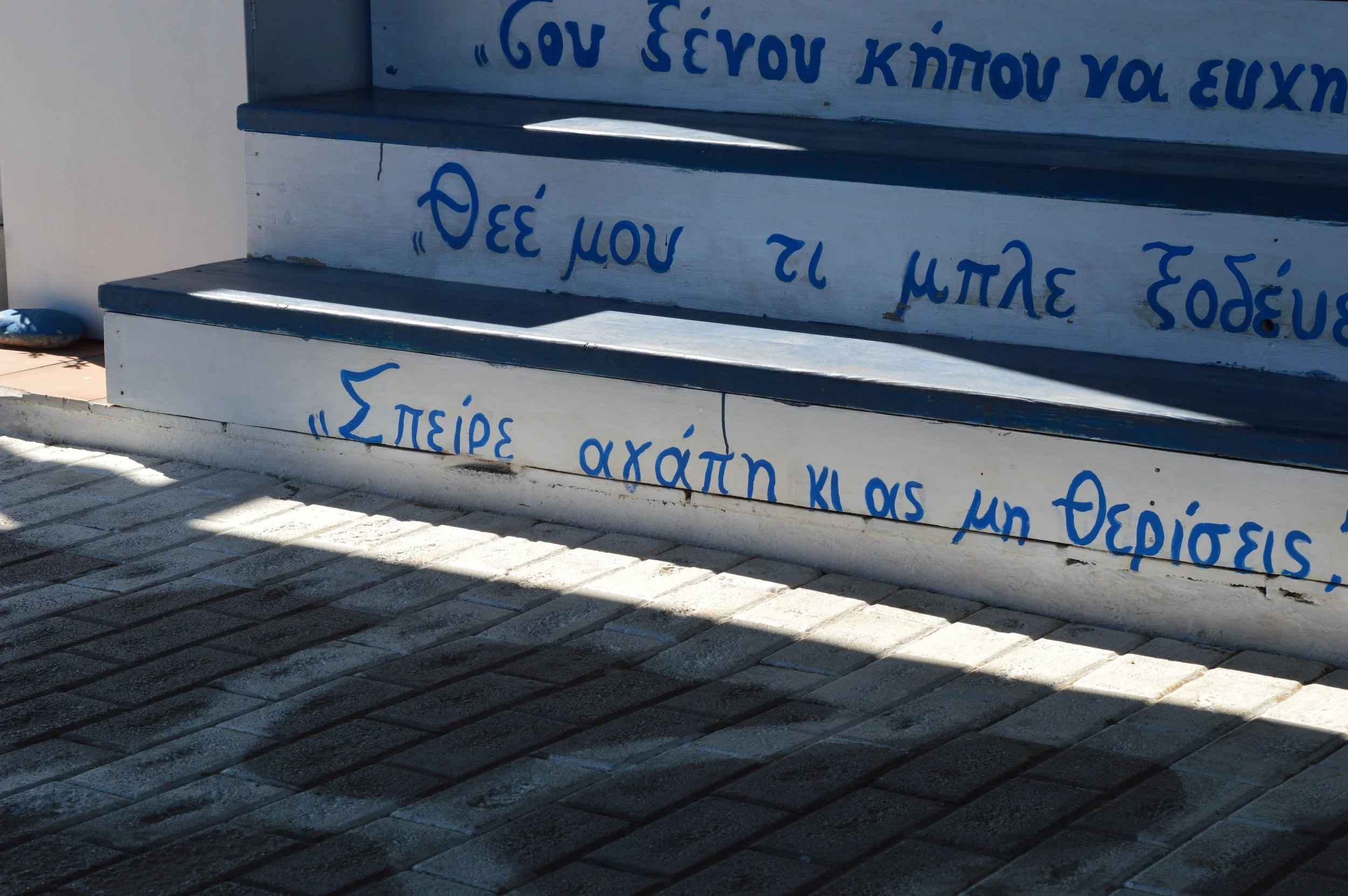 Gelateria Maria - Greek inscription painted in blue on white steps, with shadows cast on the paved ground.