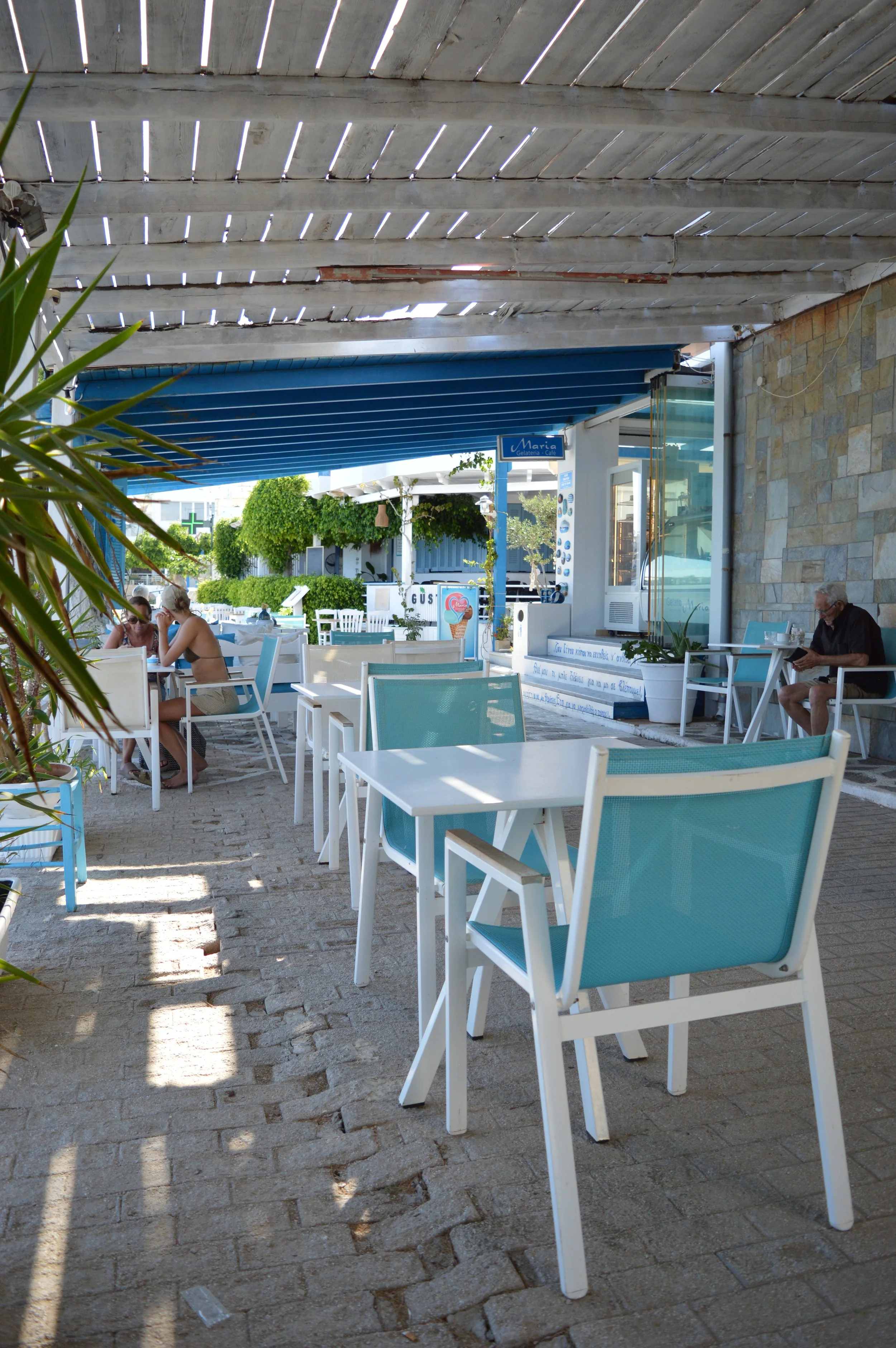 Gelateria Maria - Empty outdoor café with white tables and light blue chairs under a weathered wood roof, with a few patrons sitting and reading.