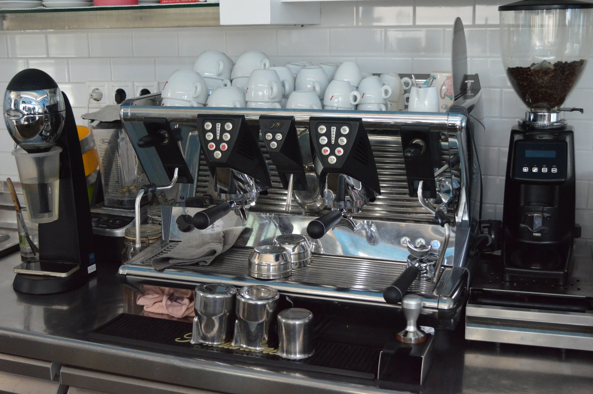 Gelateria Maria - Commercial espresso machine with portafilter handles and steam wand, cups stacked on top, coffee grinder to the right, white tiled wall in the background.