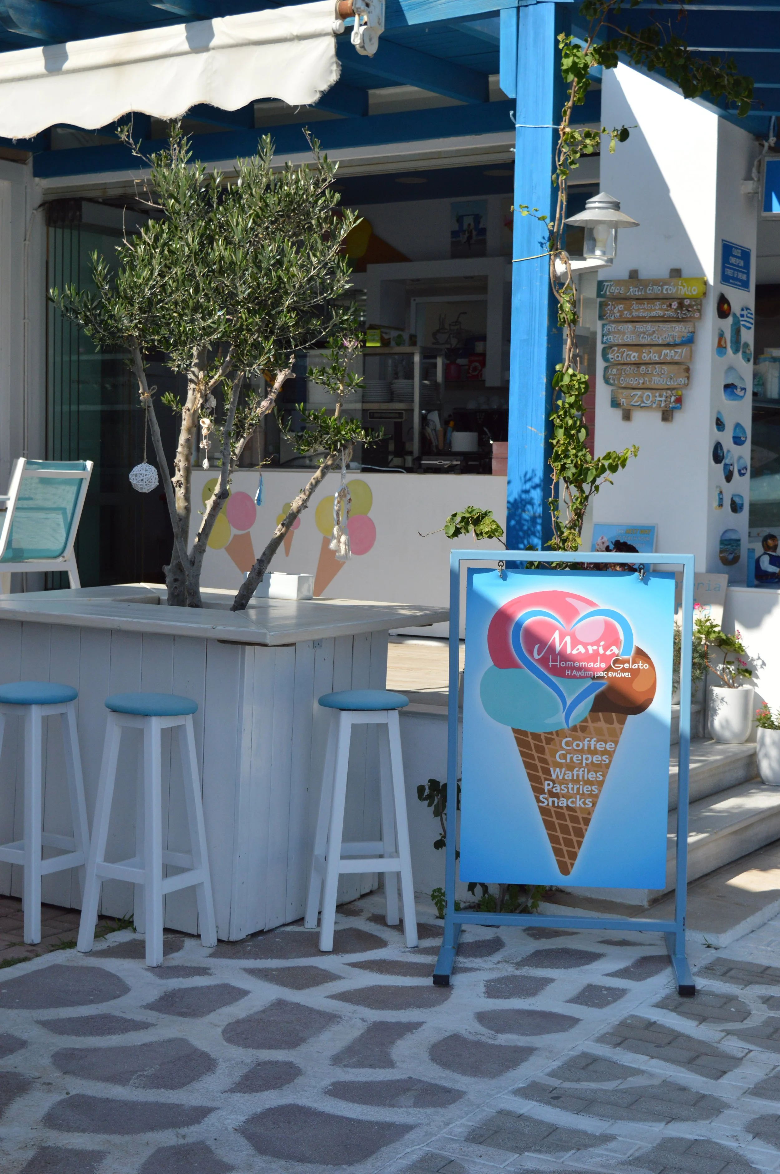 A sidewalk cafe with a white table, three white stools with blue cushions, a potted tree with hanging decorations, and a blue sign advertising homemade gelato with flavors including coffee, crepes, waffles, pastries, and snacks. The shop has a blue a