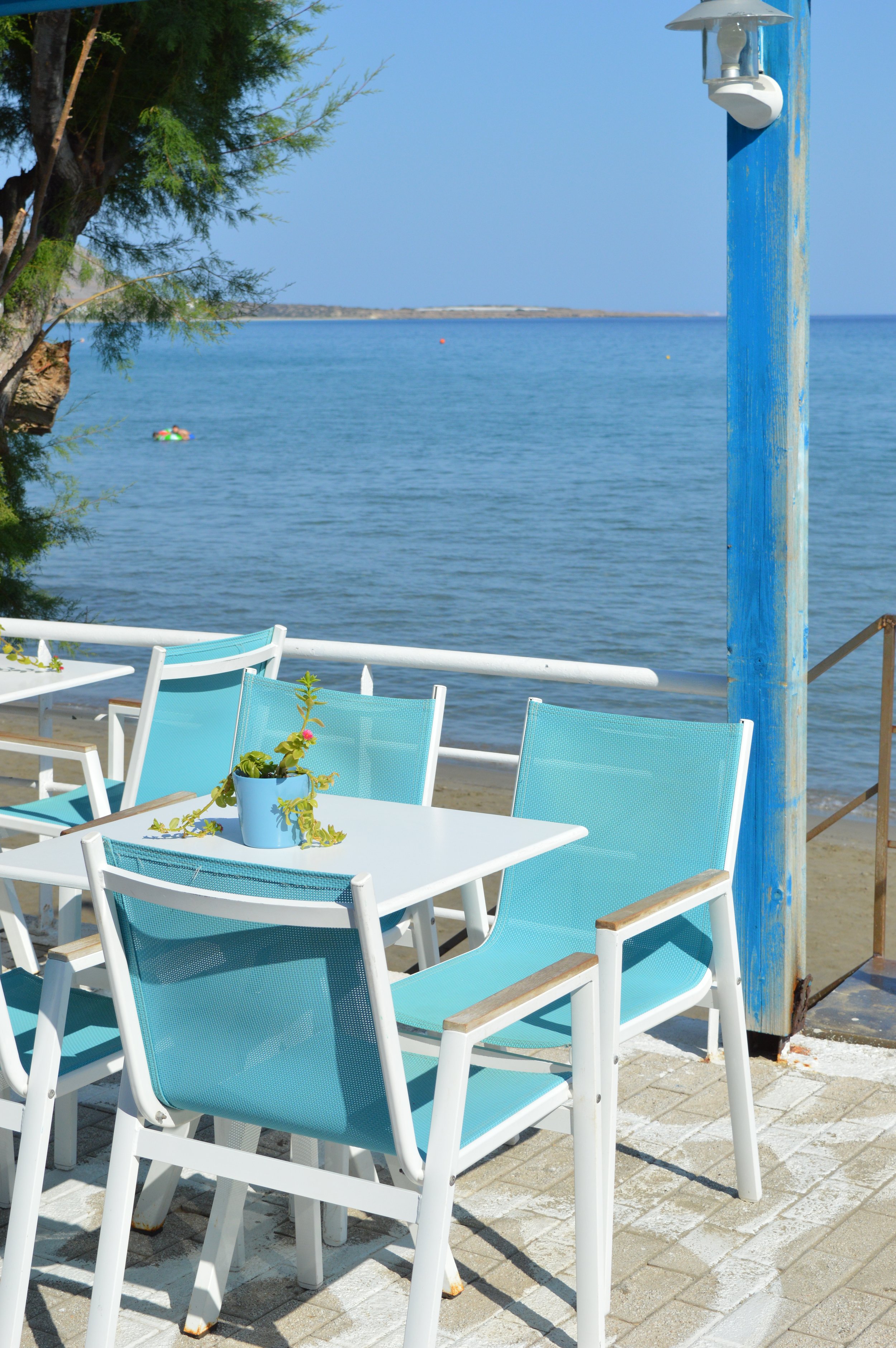 Gelateria Maria - Seaside outdoor patio with turquoise chairs around a white table, a small potted plant, overlooking the ocean with a clear blue sky.