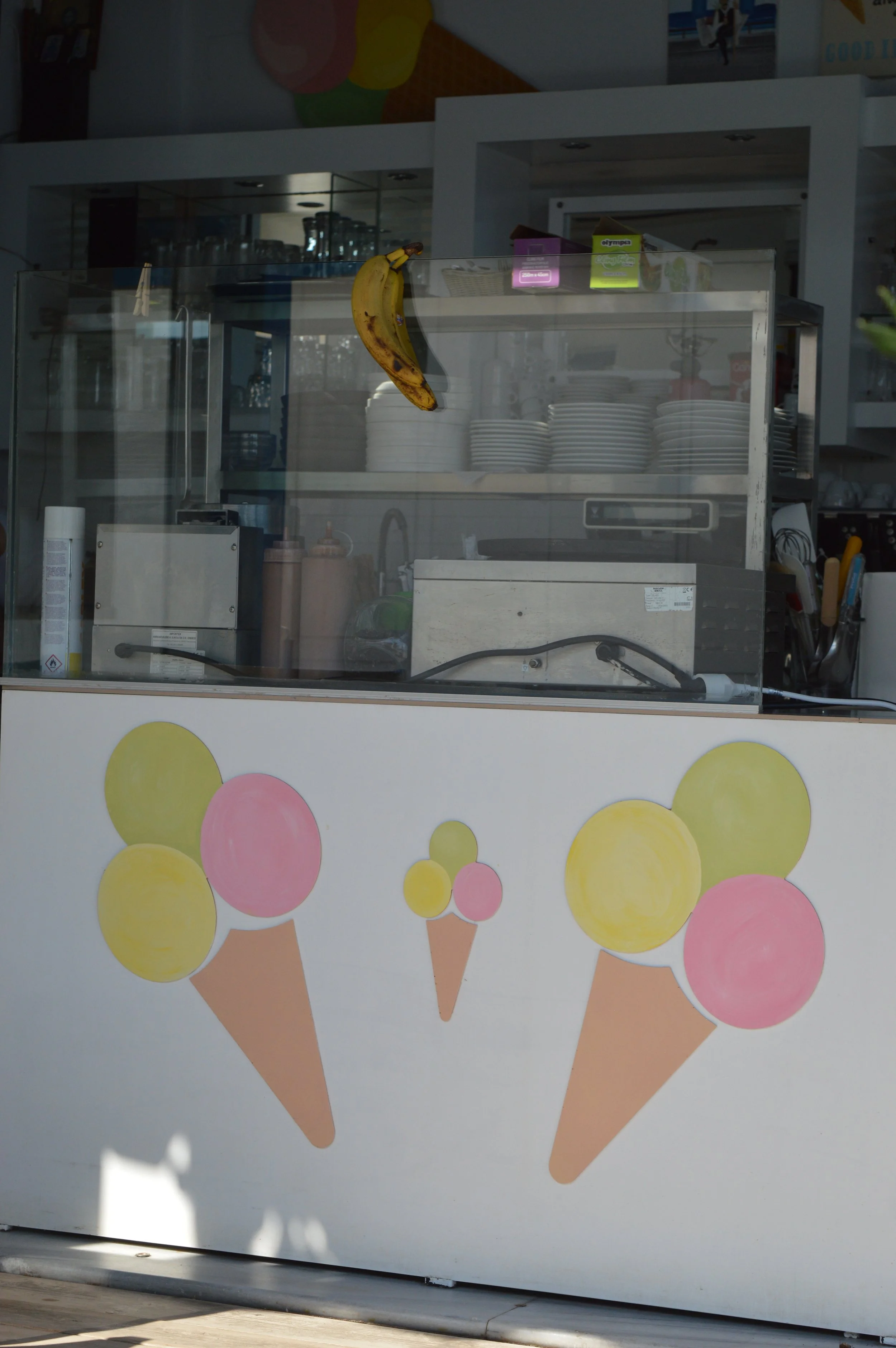 Front of an ice cream stand with a painted decoration of three ice cream cones with pastel-colored scoops on the front panel. A banana is hanging on the top of the stand.