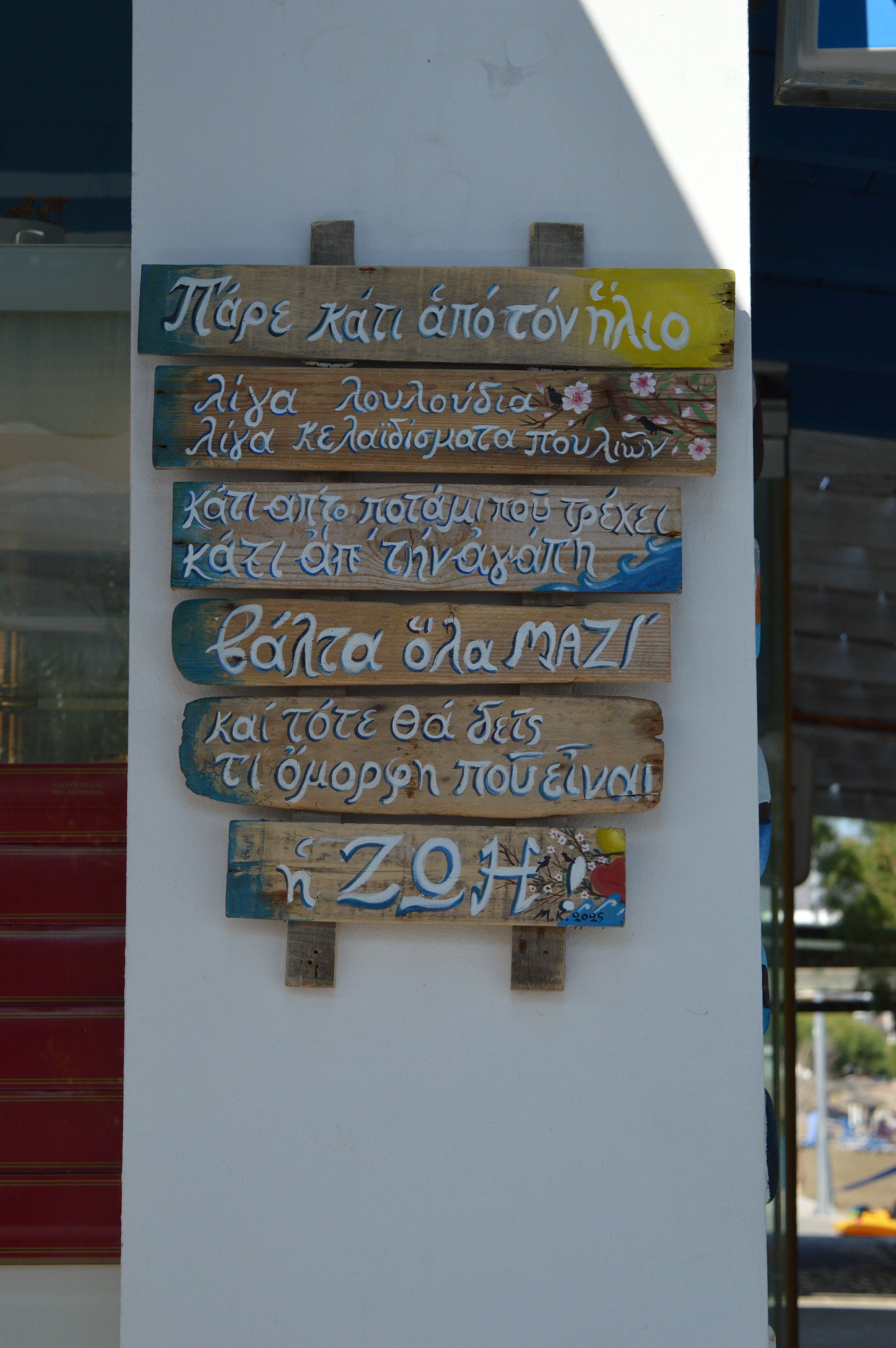 Wooden sign with Greek writing and decorative flowers hanging on a white wall outside.