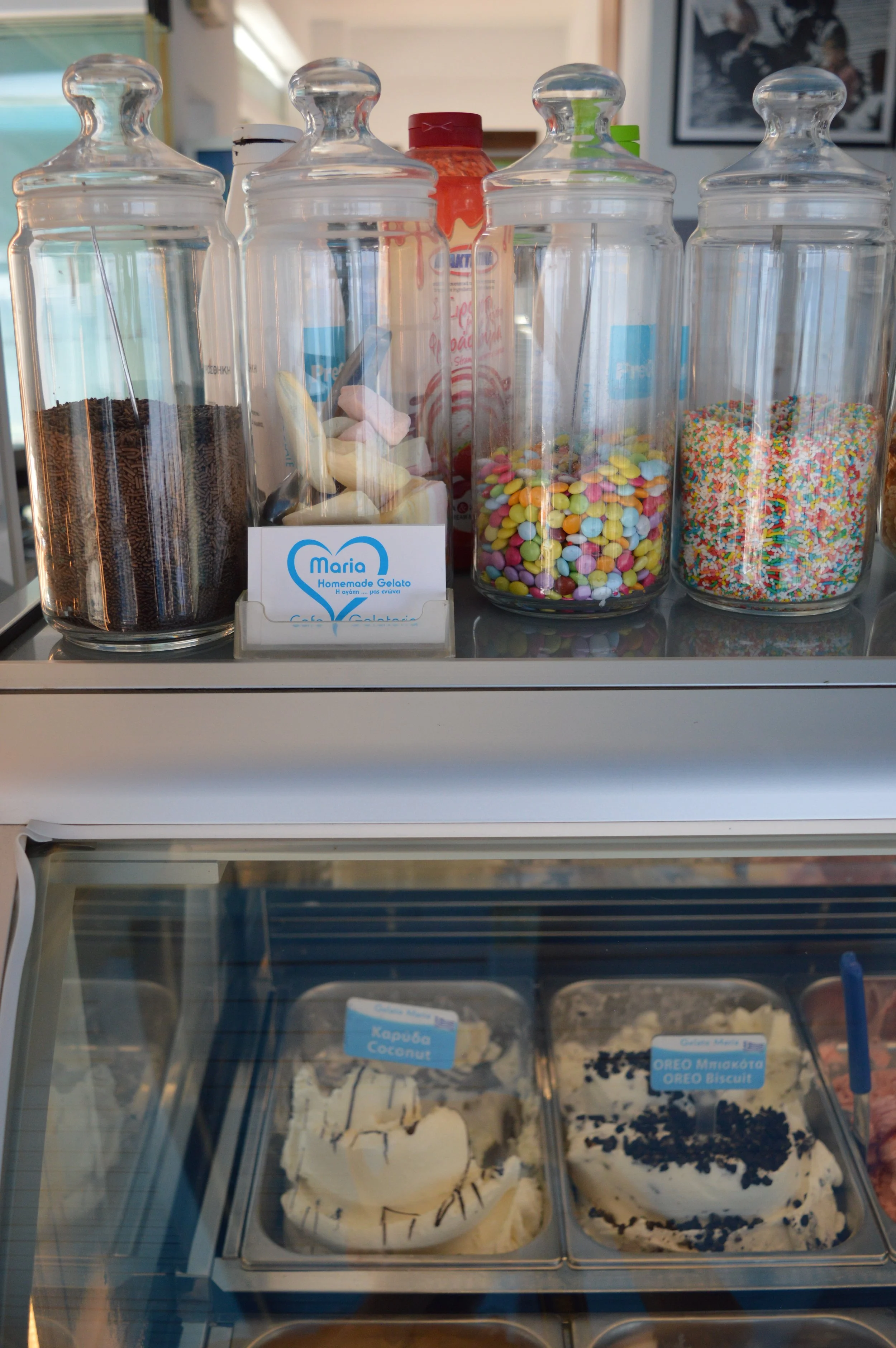 Glass jars containing sprinkles, candies, and chocolate powder on a counter, with tubs of ice cream labeled as coconut and Oreo biscuit in a refrigerated display case.