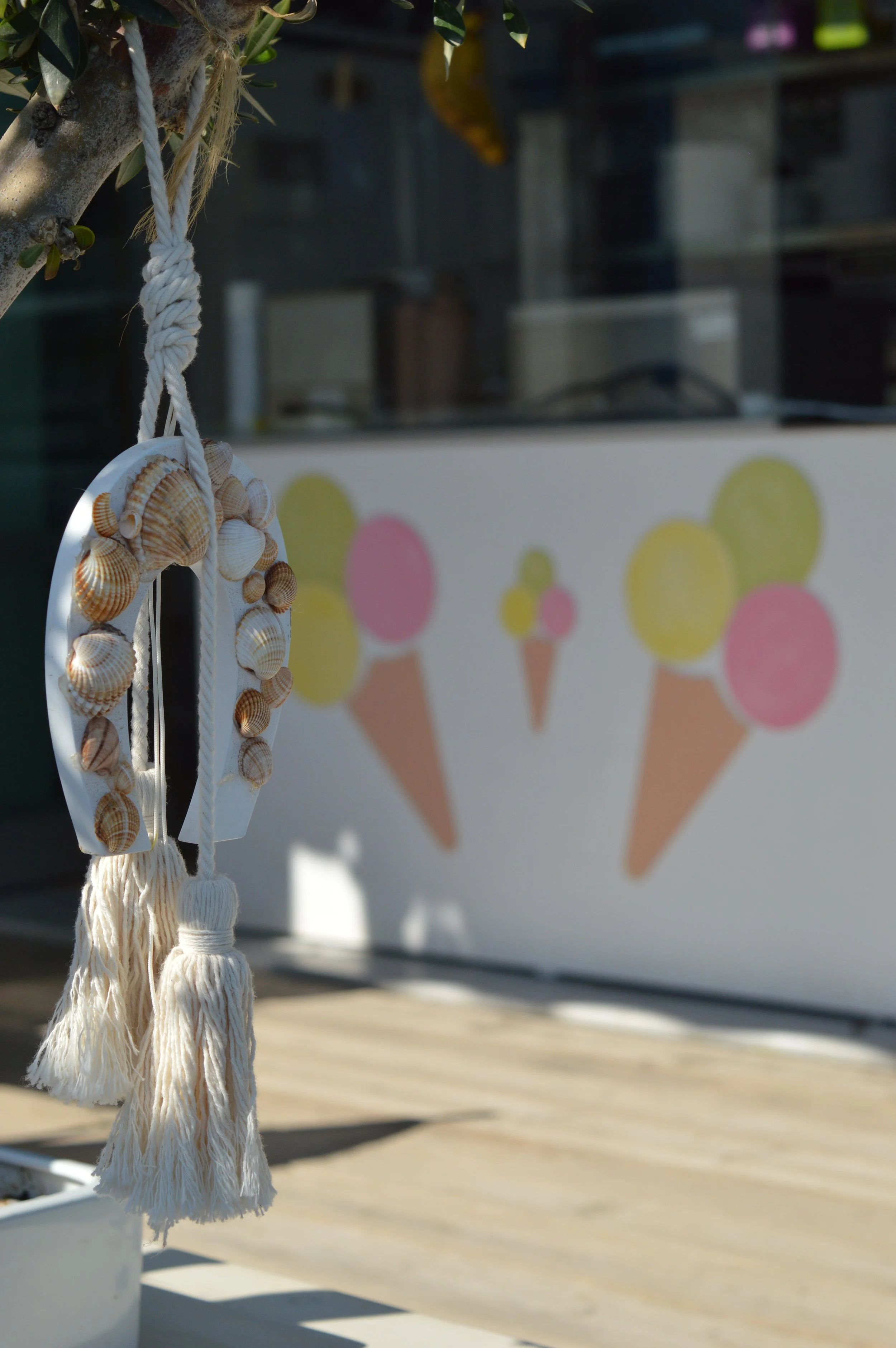Gelateria Maria - A decorative element with seashells hanging on a white rope tassel, attached to a tree. In the background, a colorful cartoon-style mural of ice cream cones with pink and yellow scoops on a white wall.