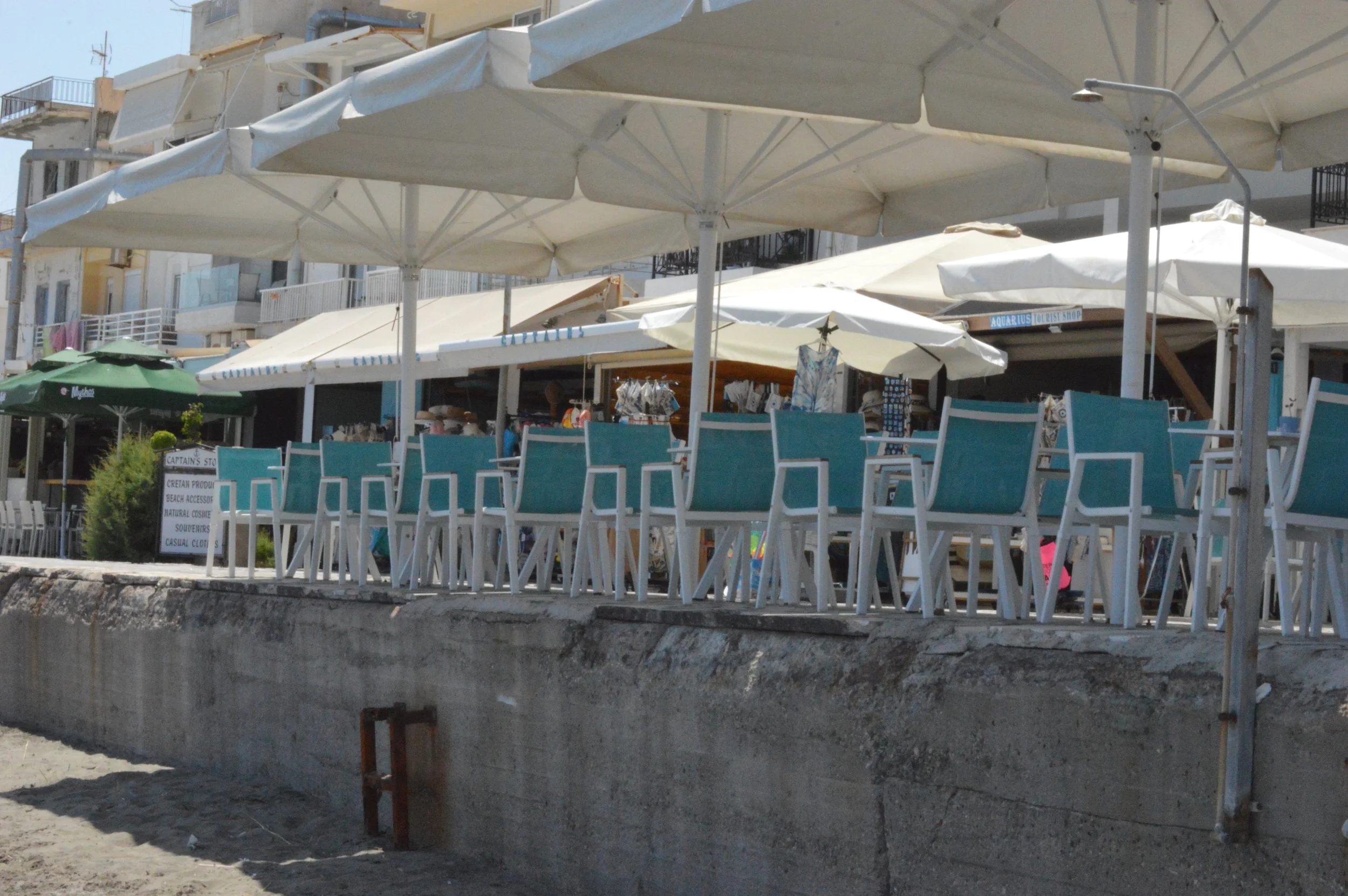 Beachside outdoor seating area with white chairs and umbrellas, near a sandy shore, with buildings and shops in the background.