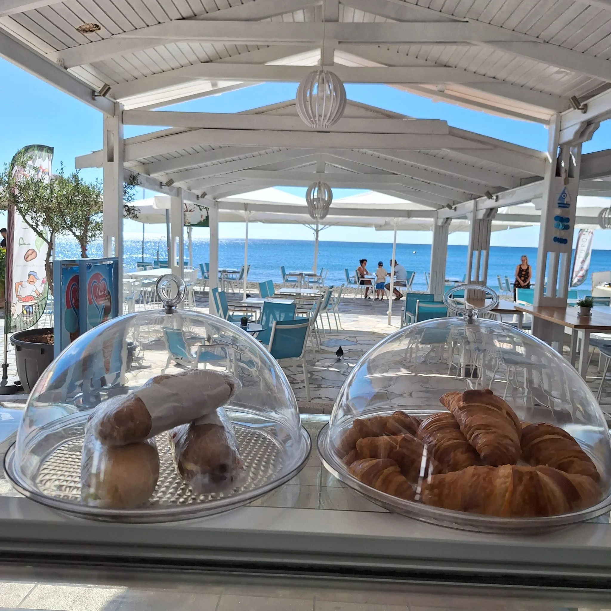 Gelateria Maria - A view from a cafe showing croissants and a wrapped pastry on display in the foreground, with a seaside patio, tables, chairs, and umbrellas under a white pergola, and the ocean and sky in the background.