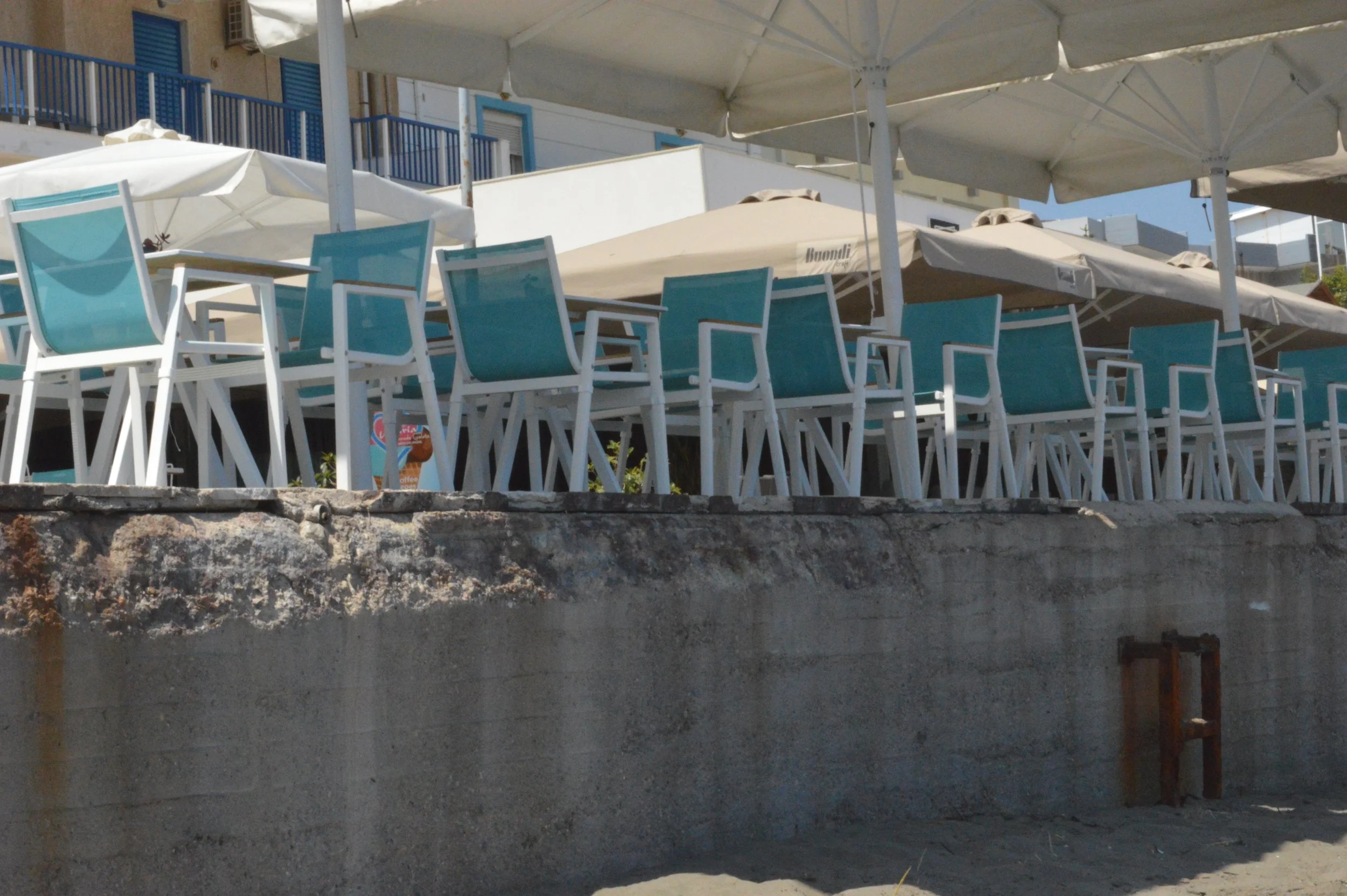 Empty outdoor seating area with turquoise chairs and large umbrellas on a patio at a beachside café or restaurant.