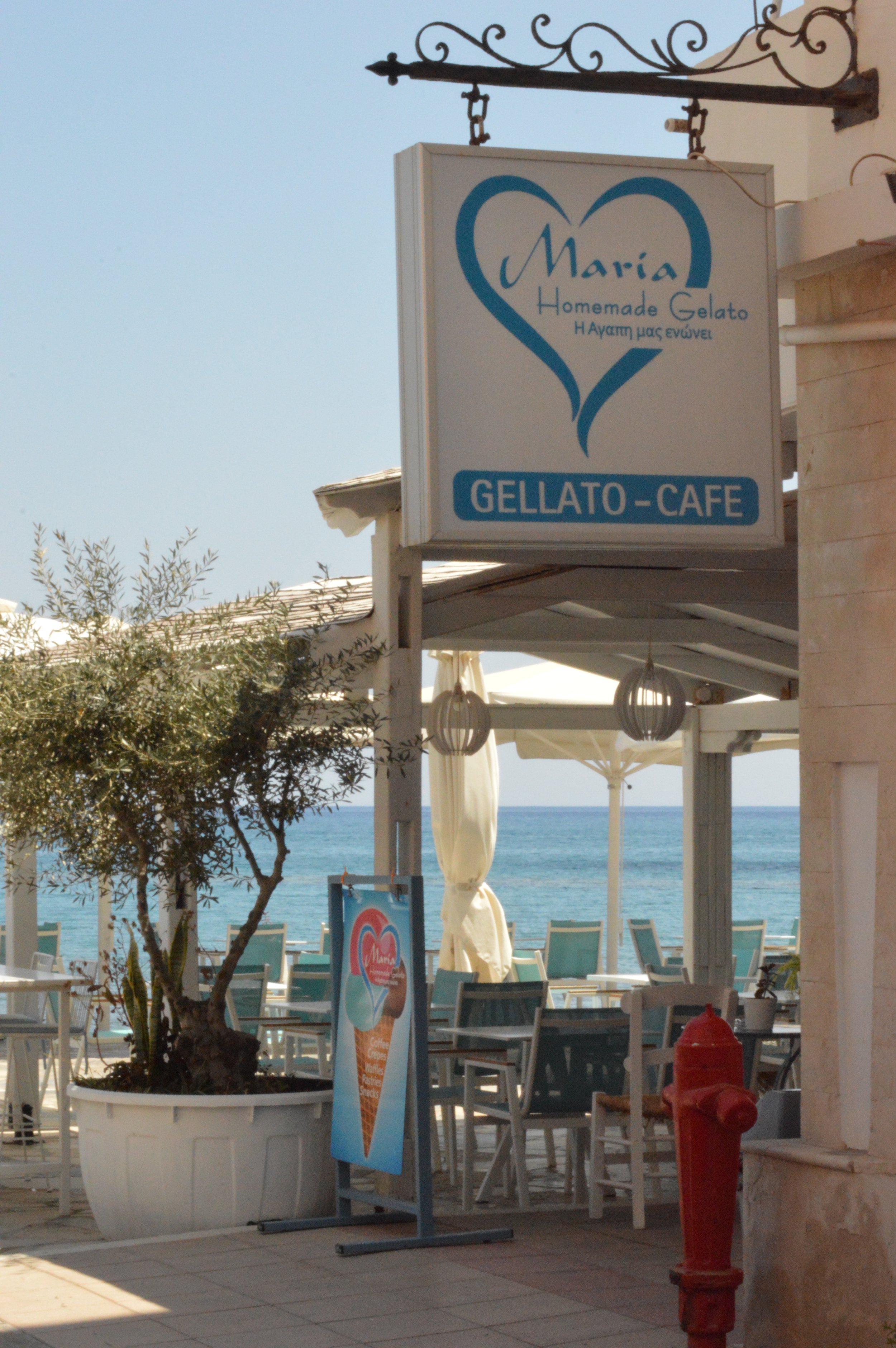 Sea view shot of an outdoor gelato café called Maria, featuring tables and chairs under umbrellas, potted trees, a signboard with an ice cream cone graphic, and the ocean in the background.