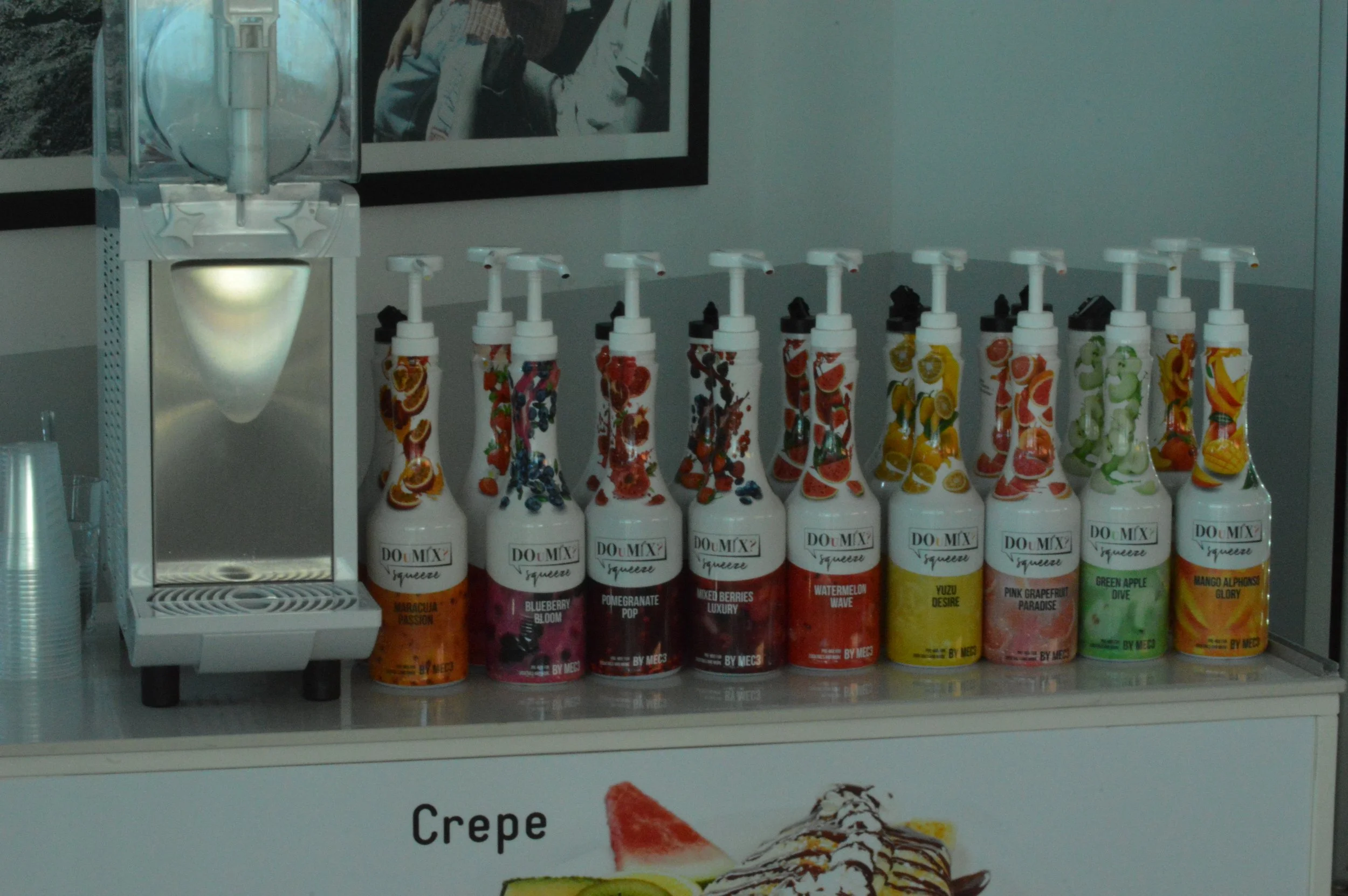 A row of colorful bottled flavored syrups with pump dispensers on a countertop, next to a beverage dispenser machine, with a partially visible advertisement featuring crepes below.