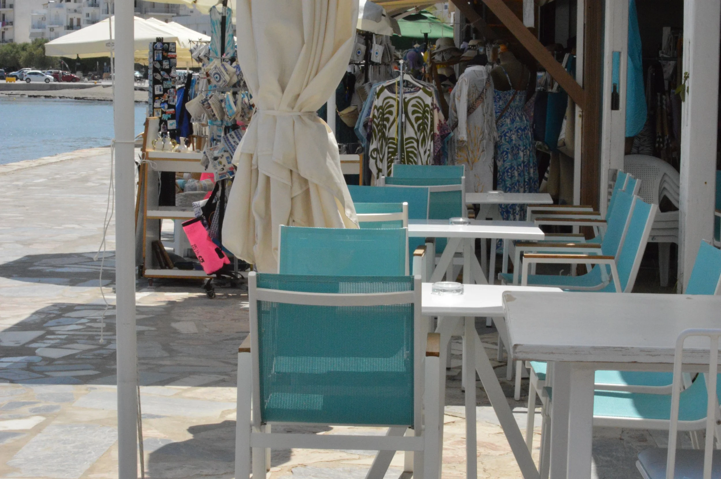 Gelateria Maria - Outdoor seating area at a seaside café with white tables and turquoise chairs, a small shop selling clothes and accessories in the background, and a view of the water and parked cars behind the scene.