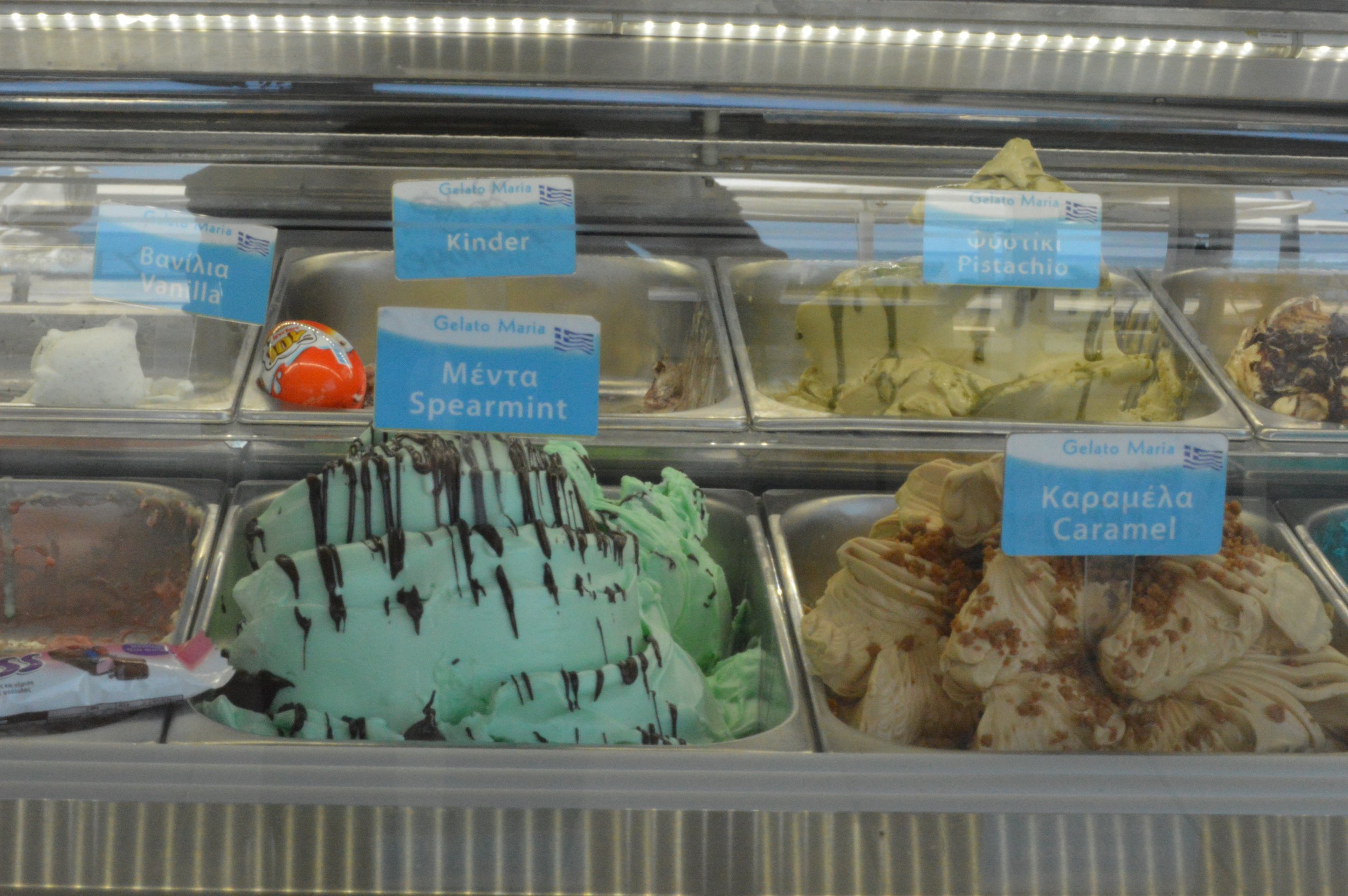 Gelateria Maria - Display case of various gelato flavors labeled in Greek and English, including spearmint, caramel, vanilla, pistachio, and others.