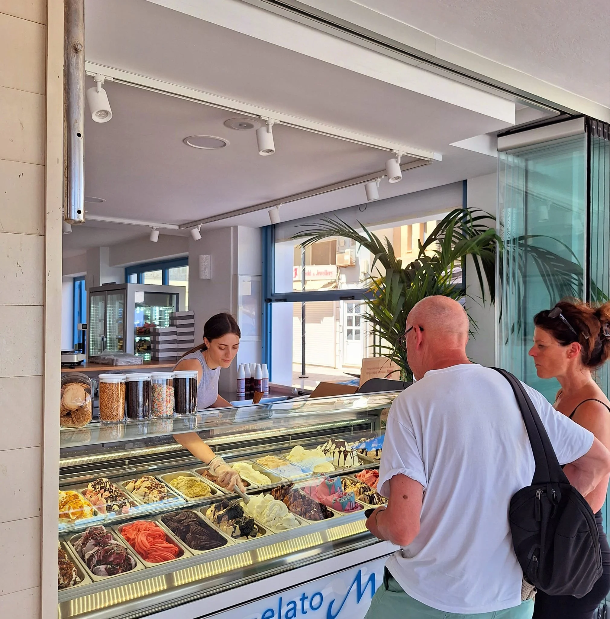 Gelateria Maria - People placing orders at an ice cream shop counter with various ice cream flavors visible inside a refrigerated display case.