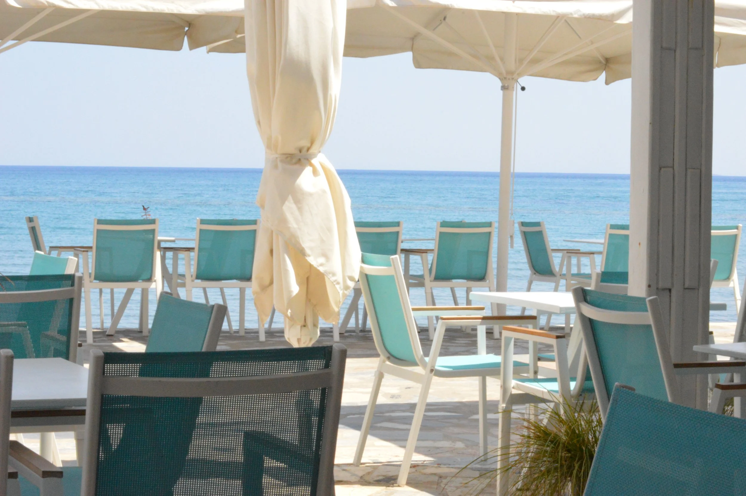Gelateria Maria - Empty outdoor beachside seating area with turquoise chairs, white tables, and large umbrellas overlooking the ocean.