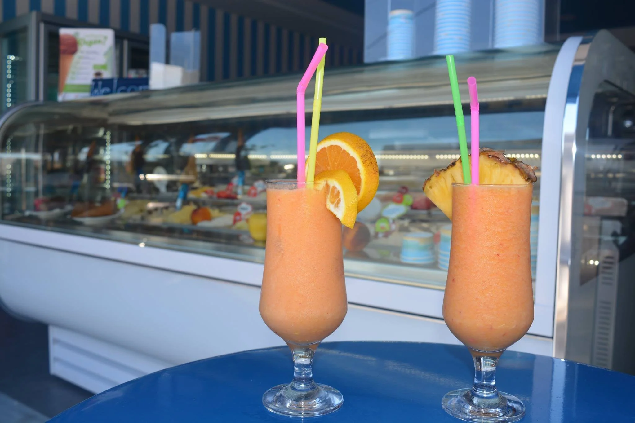 Gelateria Maria - Two tropical smoothies garnished with orange and pineapple slices on a blue table outside a gelato shop.