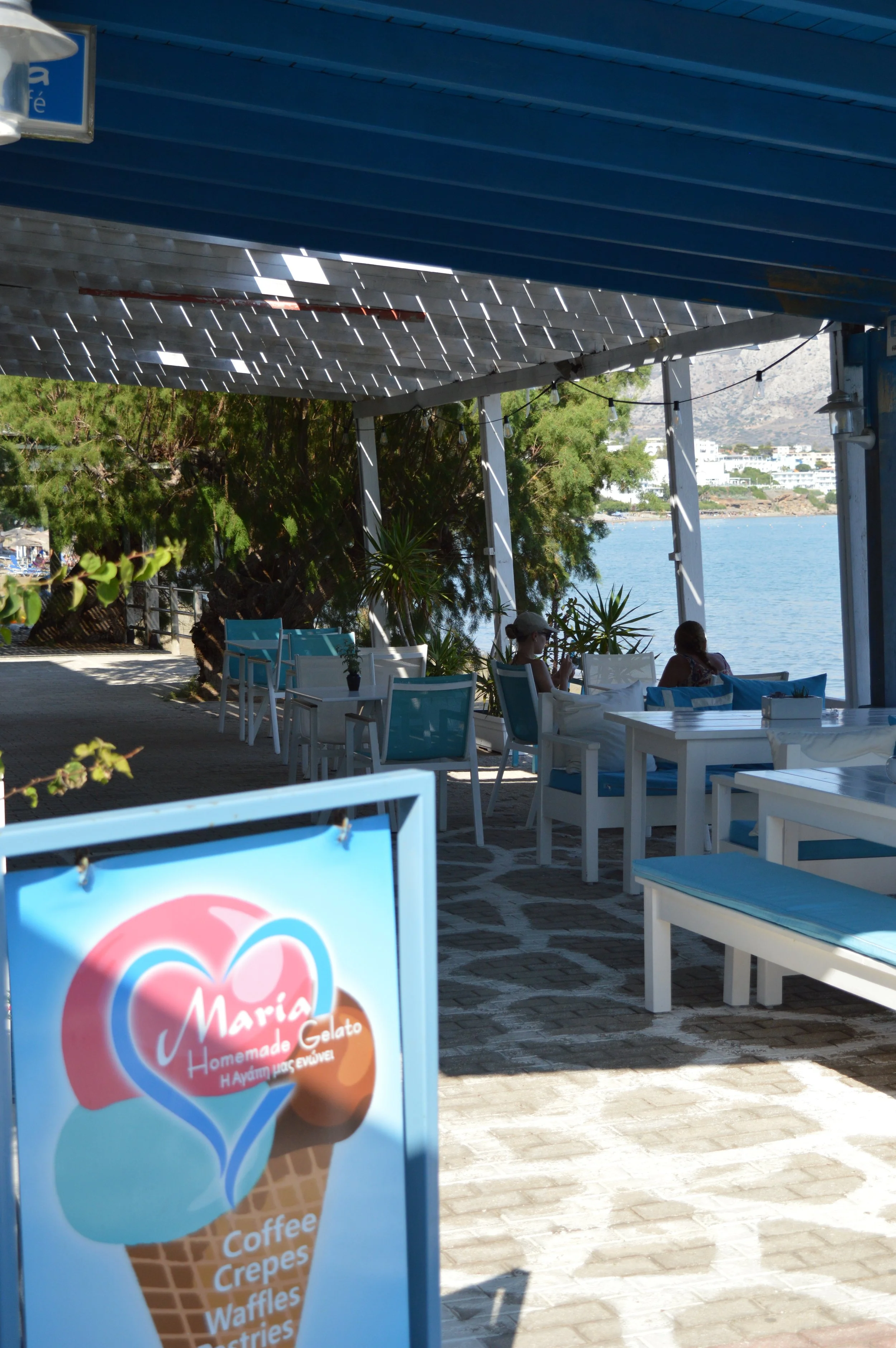 Outdoor seaside cafe with white and blue furniture, plants, and a view of the ocean in the background. A sign for Maria Homemade Gelato features an ice cream cone.