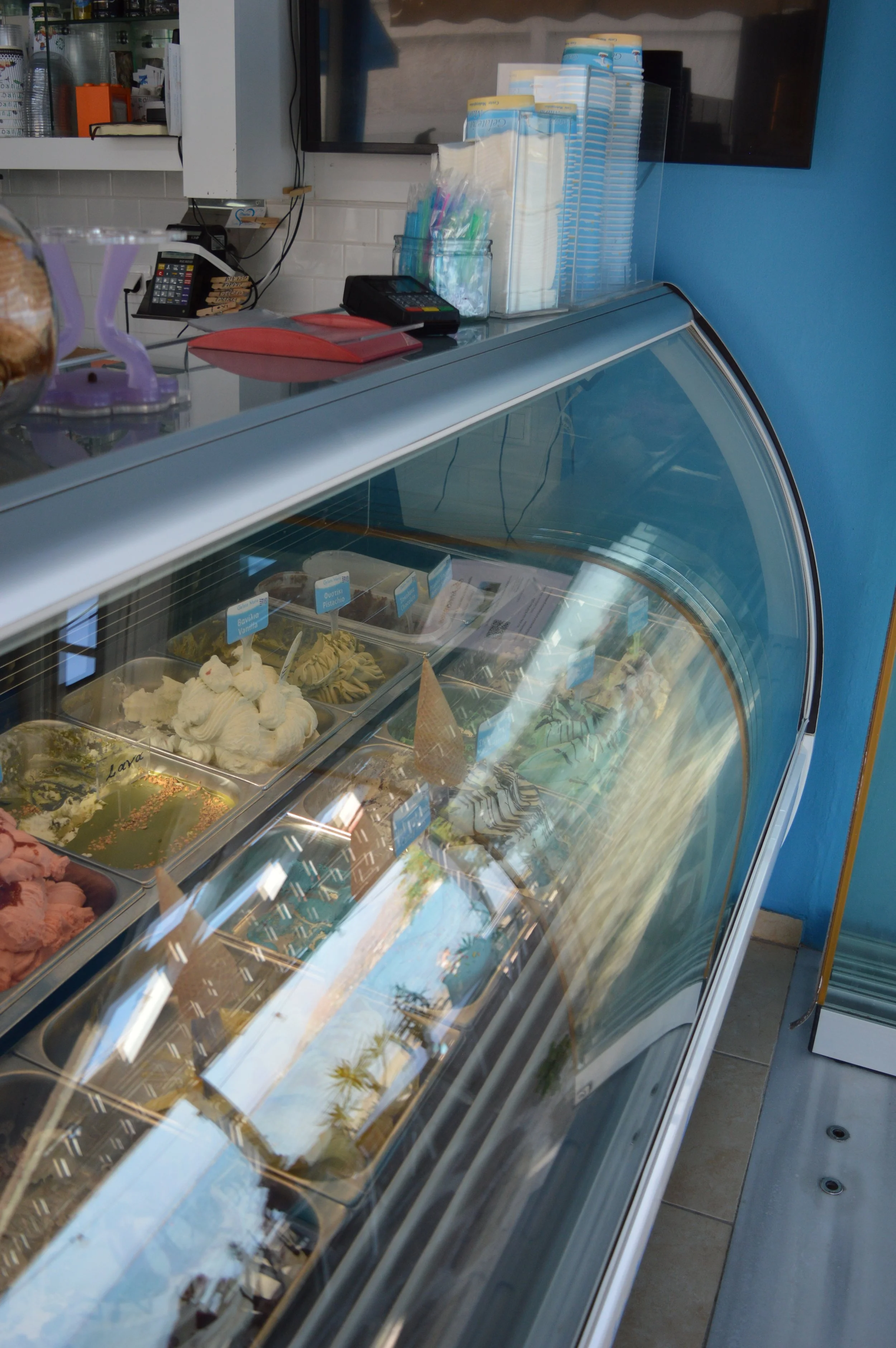 Gelateria Maria - Ice cream display case in an ice cream shop with various flavors, behind a glass top and with a reflection of sunny outdoor trees.