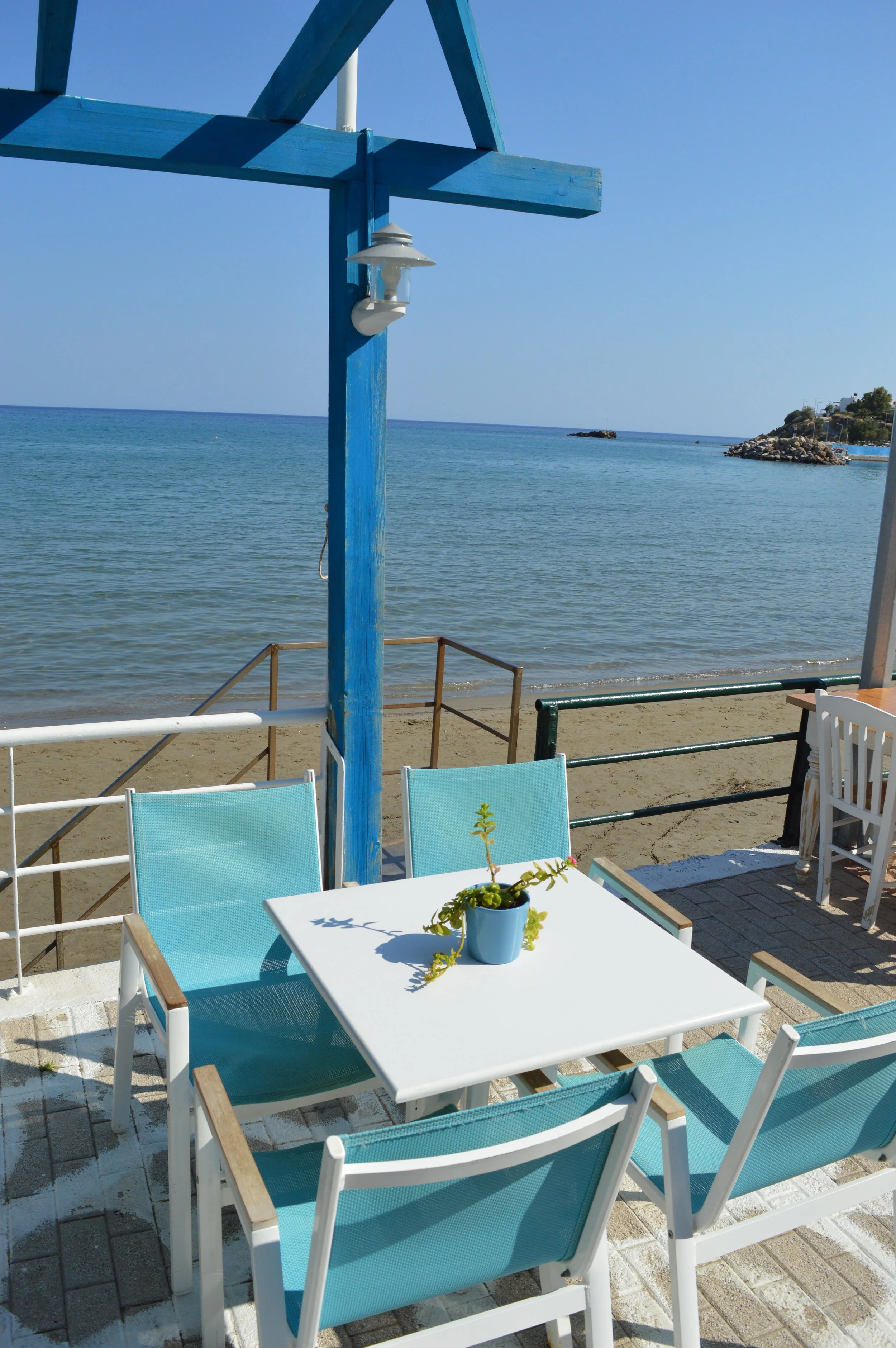 Beachside outdoor seating with a white table and turquoise chairs, overlooking the ocean with a sandy beach, rocks, and a clear blue sky.