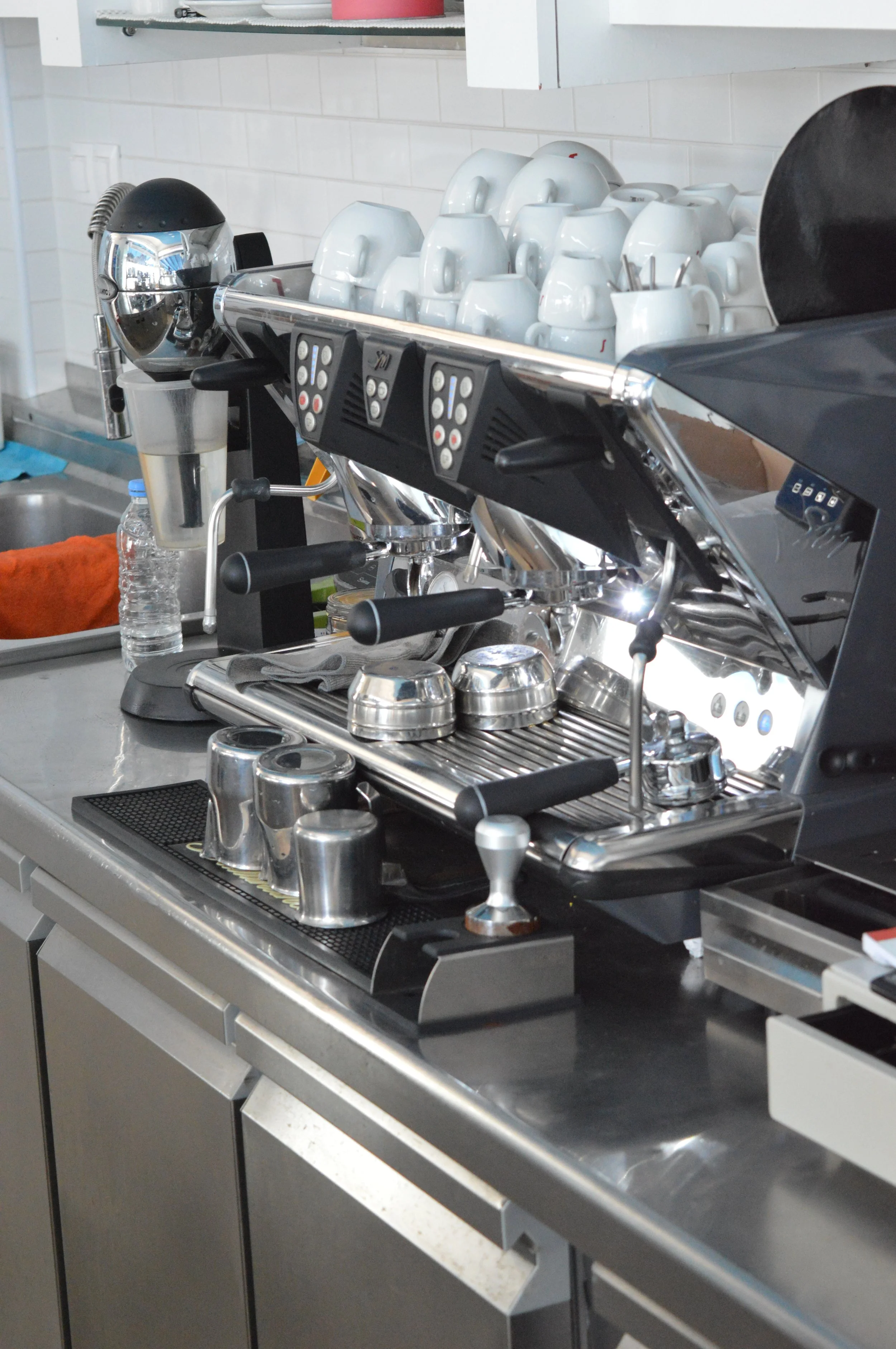 Gelateria Maria - Commercial espresso machine with cups, grinder, and water bottles on a stainless steel countertop in a coffee shop kitchen.