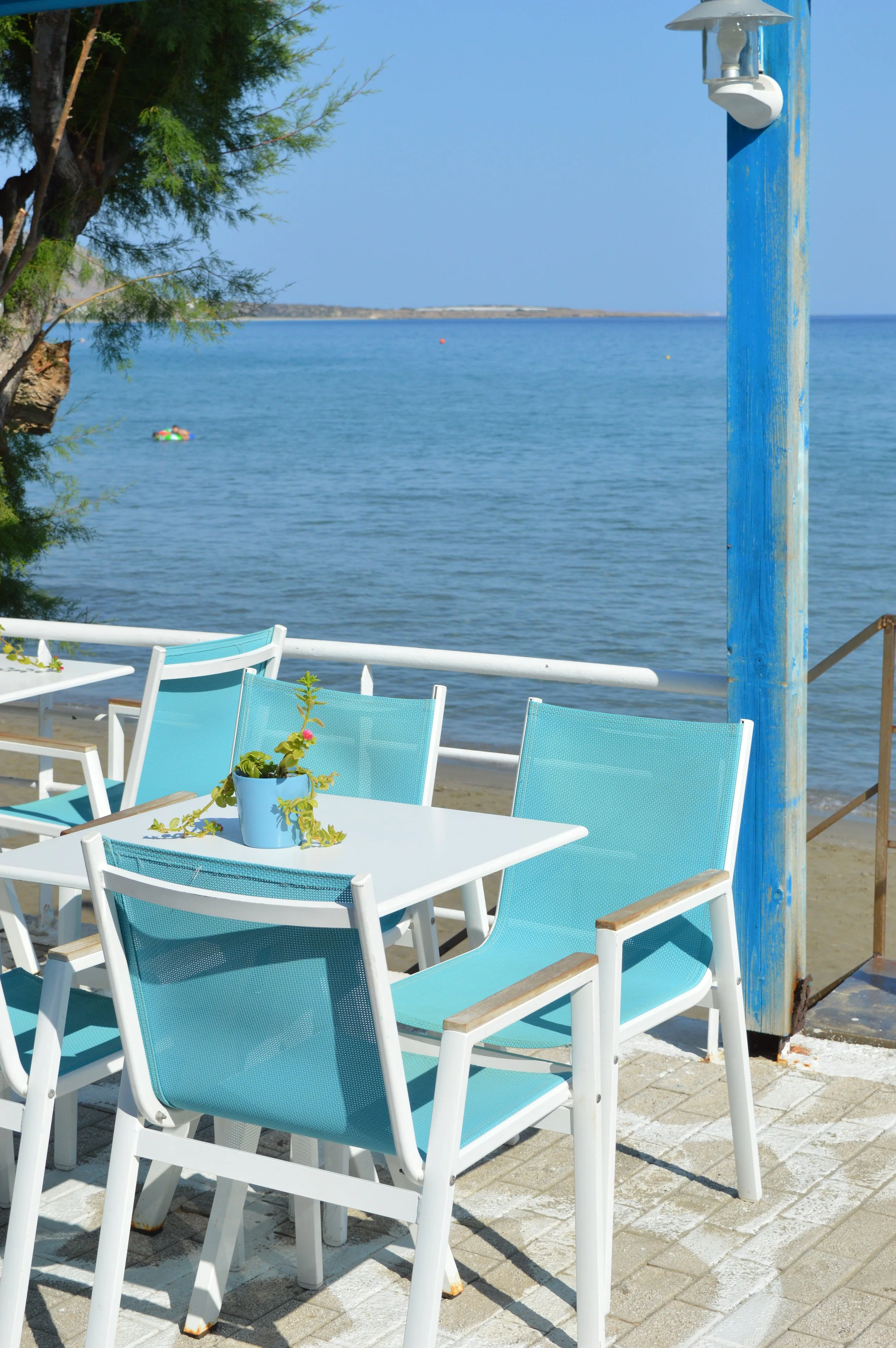 Gelateria Maria - Outdoor patio near the beach with a white table, several blue chairs, a potted plant on the table, and a view of the ocean in the background.