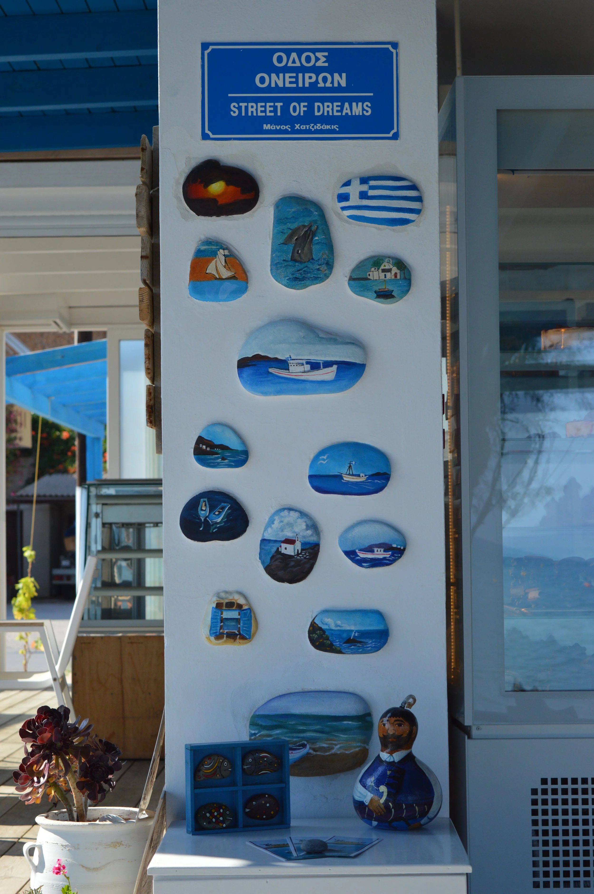Decorative wall with painted dishes and a sign that reads 'Street of Dreams' in English and Greek. The dishes depict coastal scenes, boats, and water, reflecting Greek island themes. A small pot with a succulent plant and a painted clay figure are at