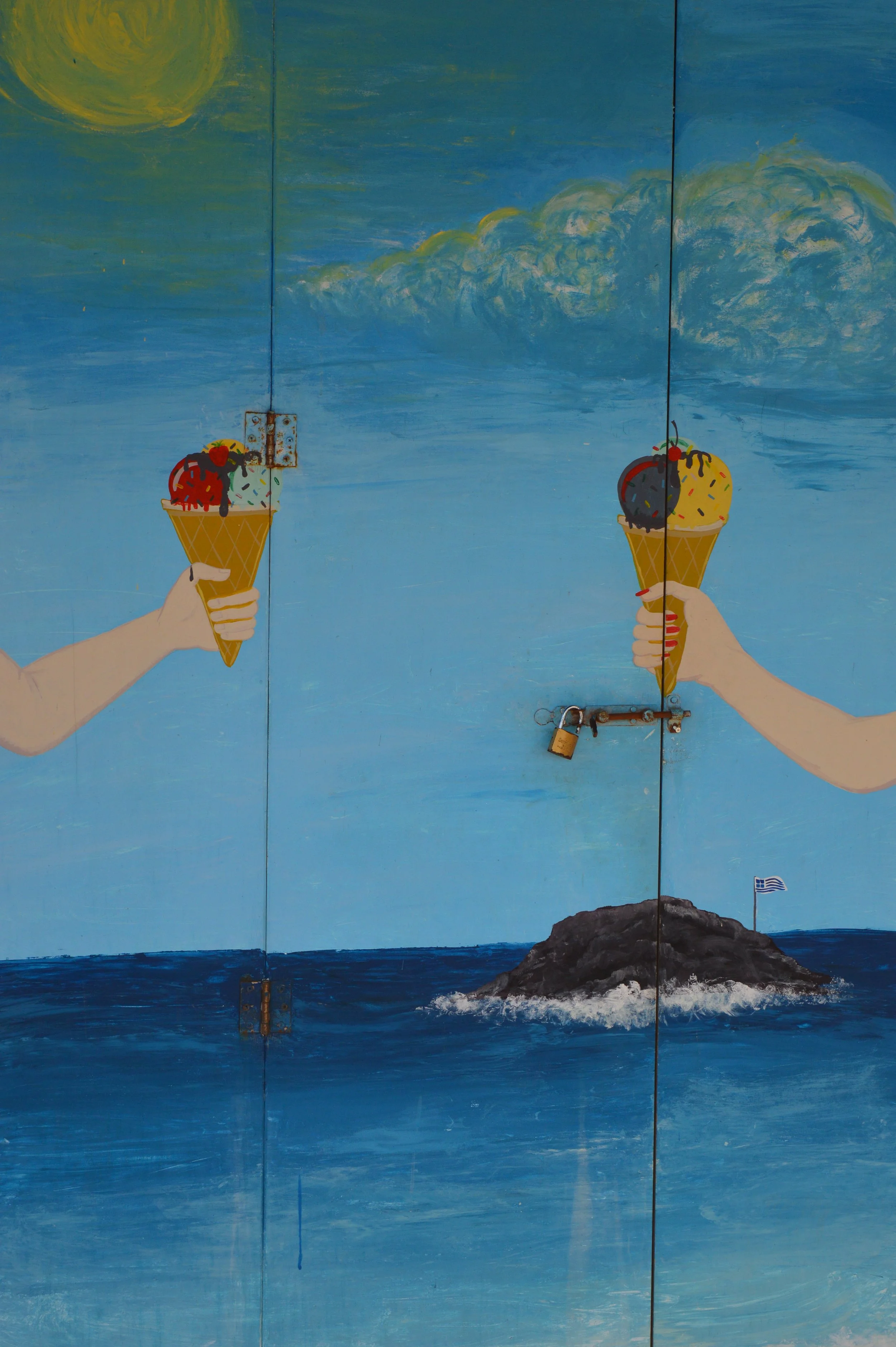 A mural painted on a blue background depicts two hands holding ice cream cones, with the ocean and a rocky island with a small Greek flag in the background. One cone has three scoops of ice cream with fruit toppings, and the other has two scoops, wit