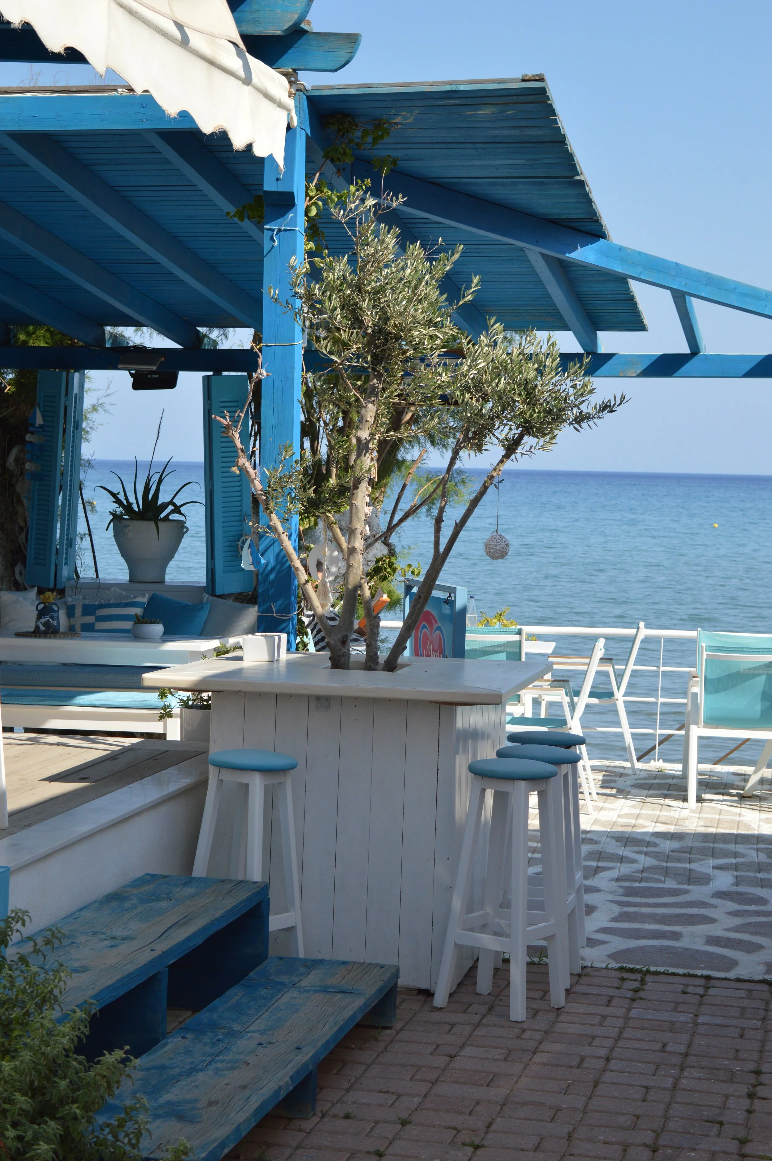 A seaside outdoor patio with white and blue furniture, a potted plant, and the ocean in the background.