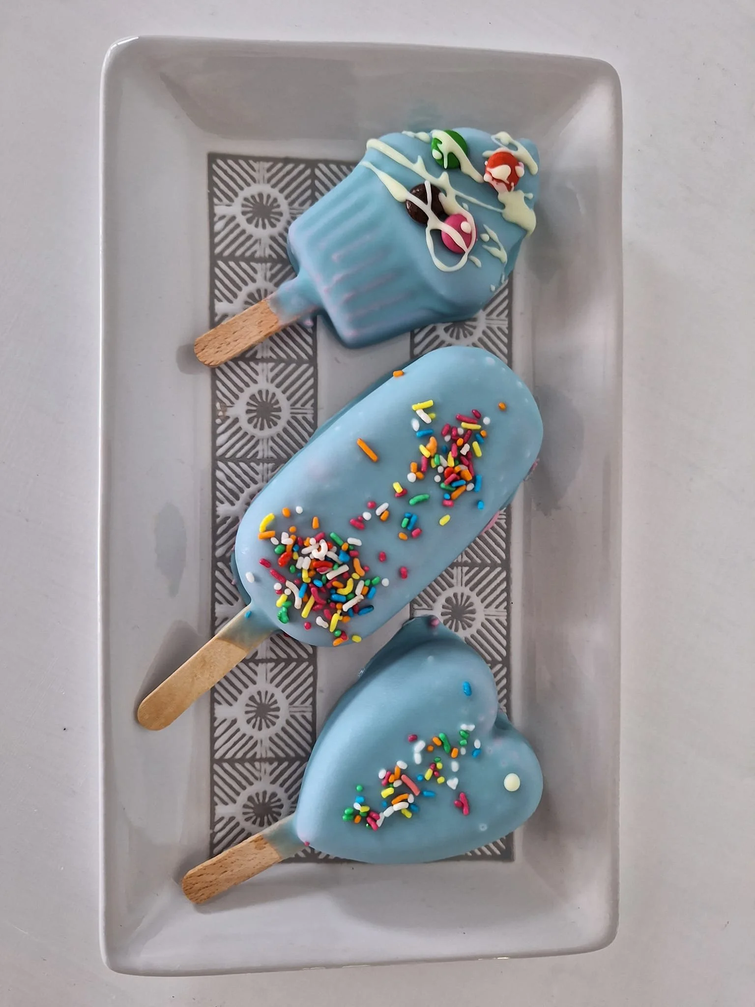 Gelateria Maria - Three blue ice cream bars with colorful sprinkles and white chocolate drizzle on a white rectangular tray.