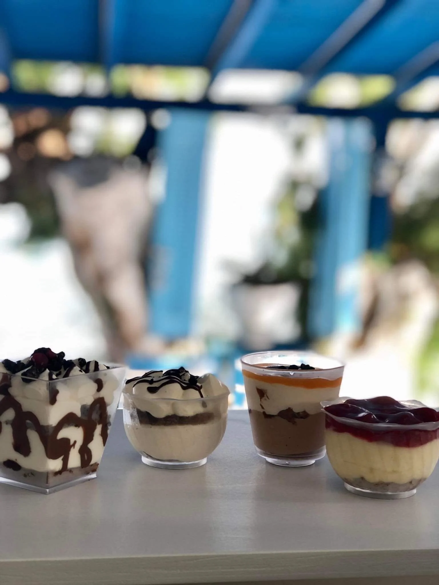 Gelateria Maria - Four bowls of dessert on a white table with a blurry outdoor background, featuring blue structure and potted plants.
