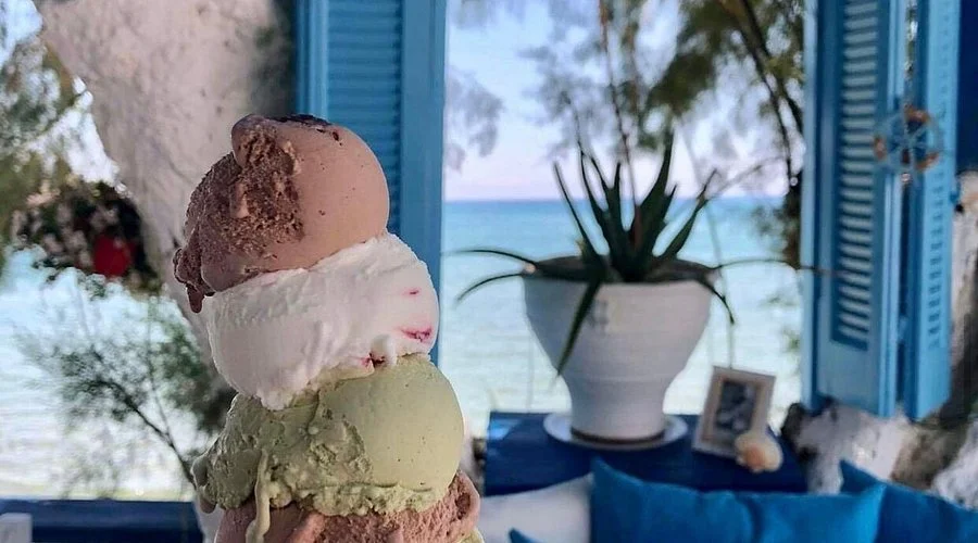 Gelateria Maria - Scoops of ice cream in front of a window with blue shutters, with a view of the ocean, a potted plant, and framed photo.