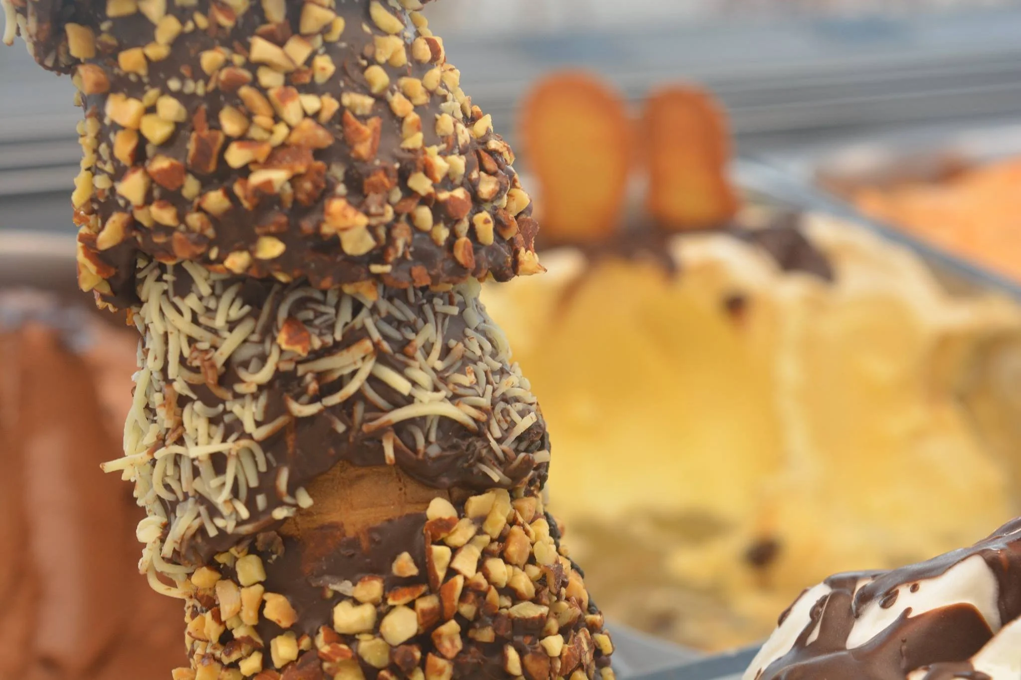 Gelateria Maria - Close-up of assorted ice cream bars coated with nuts, chocolate, and sprinkles, with ice cream and more treats blurred in the background.