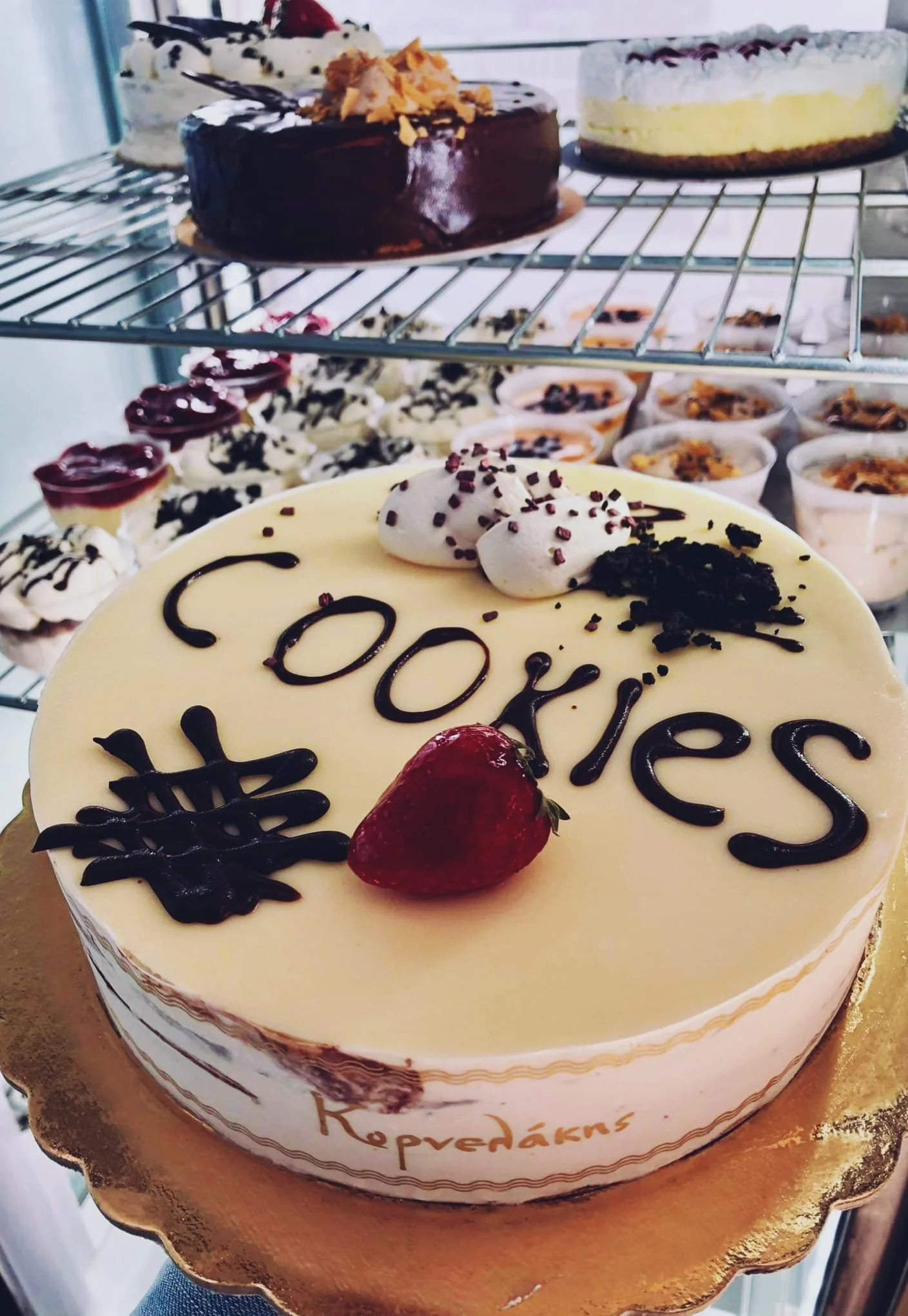 Gelateria Maria - A birthday cake with a strawberry and the word 'cookies' written on top in chocolate, surrounded by whipped cream, sprinkles, and chocolate decorations. In the background are multiple other cakes and desserts on display.