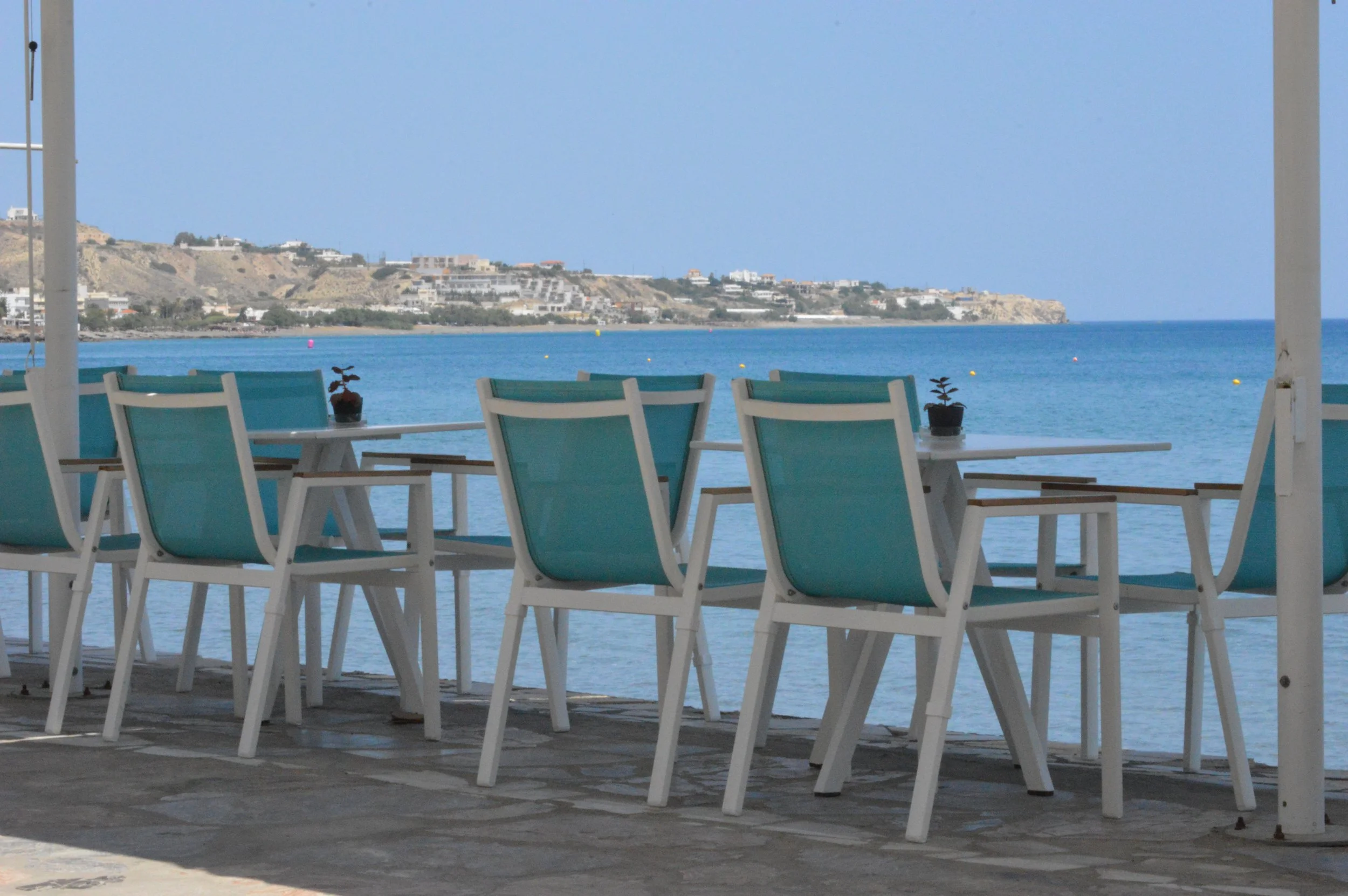 Gelateria Maria - Empty outdoor seating area with turquoise chairs and white tables, overlooking the ocean with a distant shoreline and hillside.