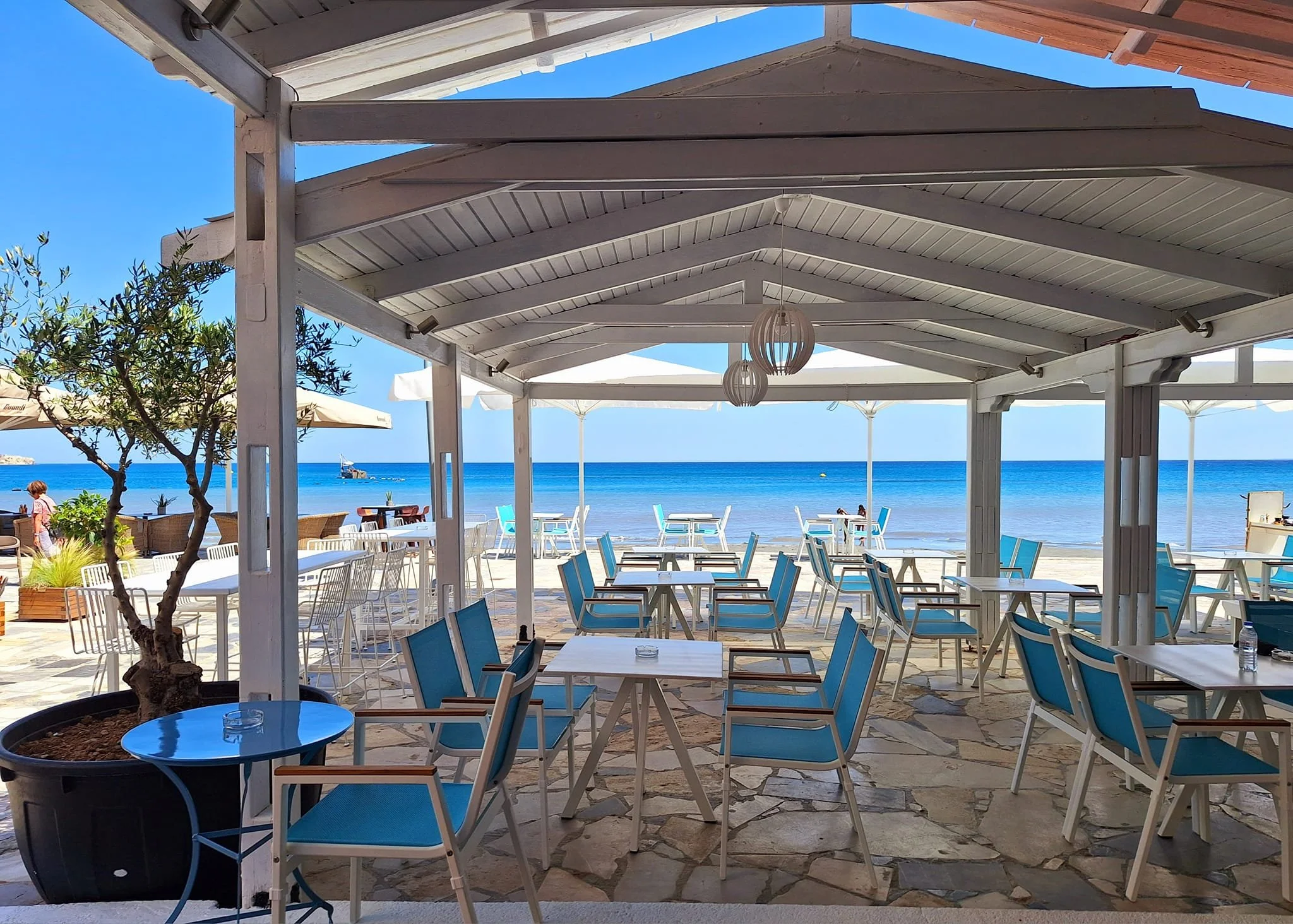 Gelateria Maria - Empty outdoor beachside restaurant with white tables and blue chairs under a white pavilion, with the ocean and boats in the background.