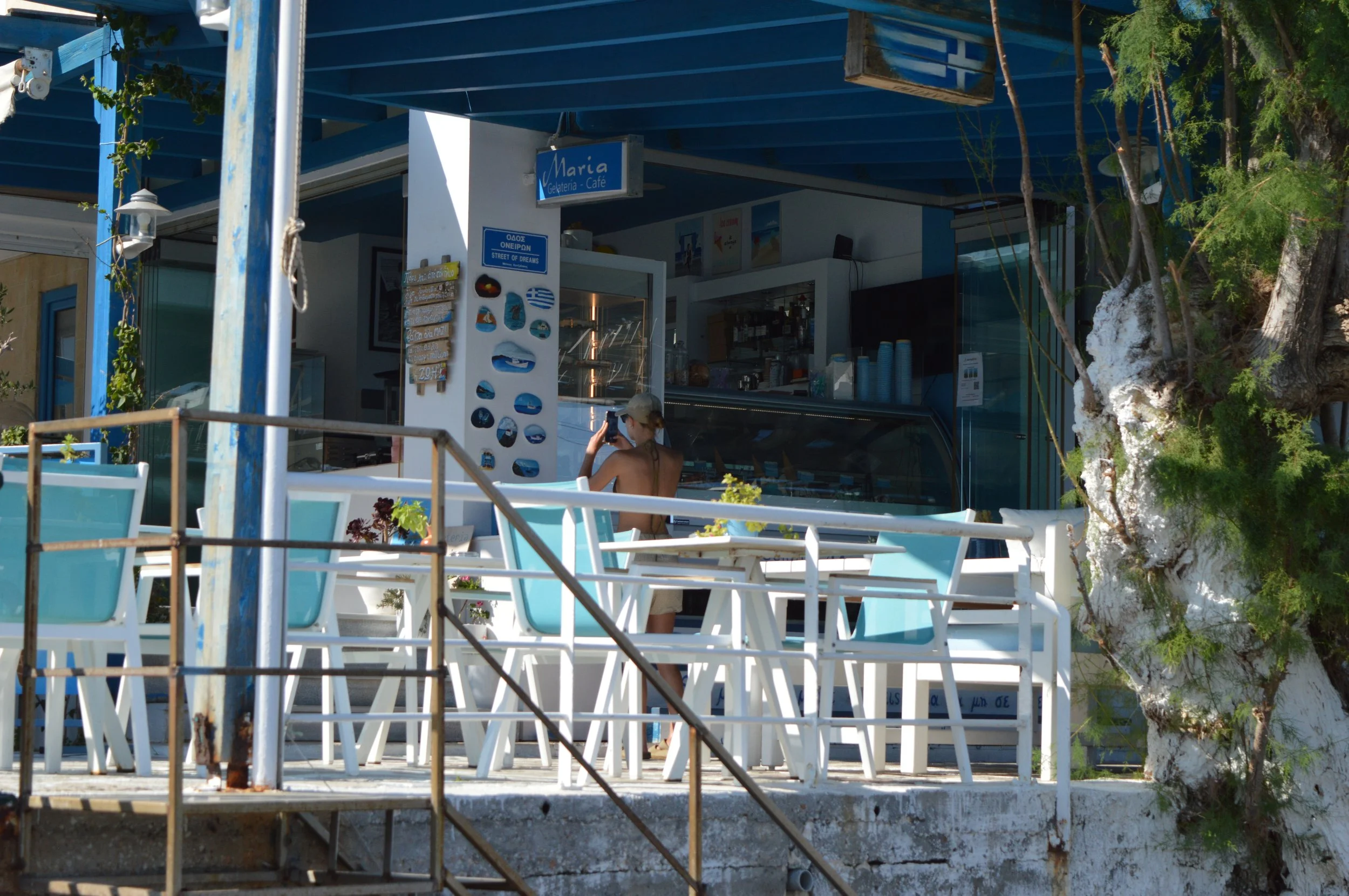 Gelateria Maria - A woman sitting outside a blue and white cafe named 'Maria' on a sunny day, taking a photo with her phone. The cafe has white tables and chairs, some potted plants, and is surrounded by green trees.