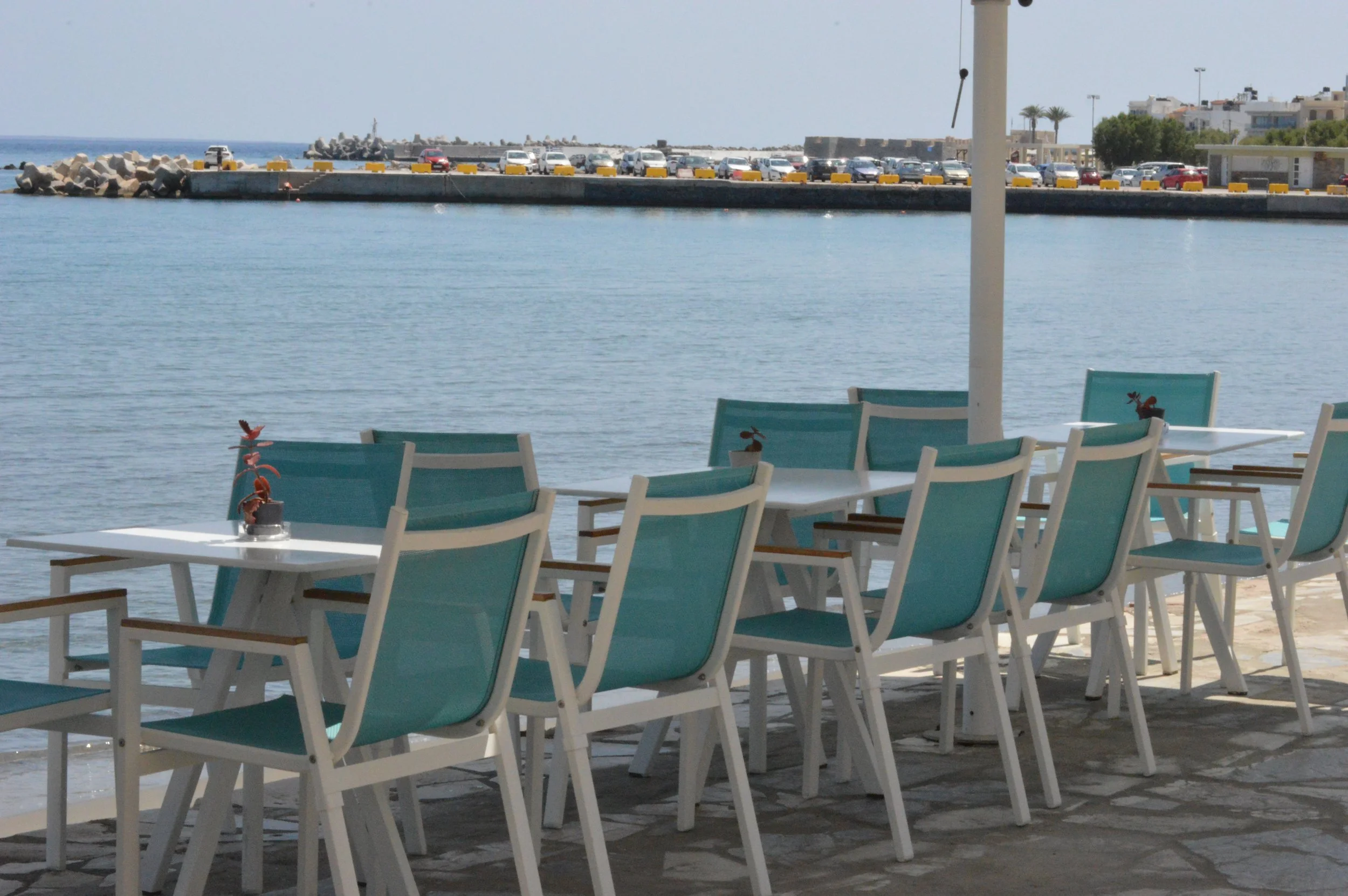Gelateria Maria - Outdoor seating area with turquoise chairs and white tables near the water, with a harbor and parked cars in the background.