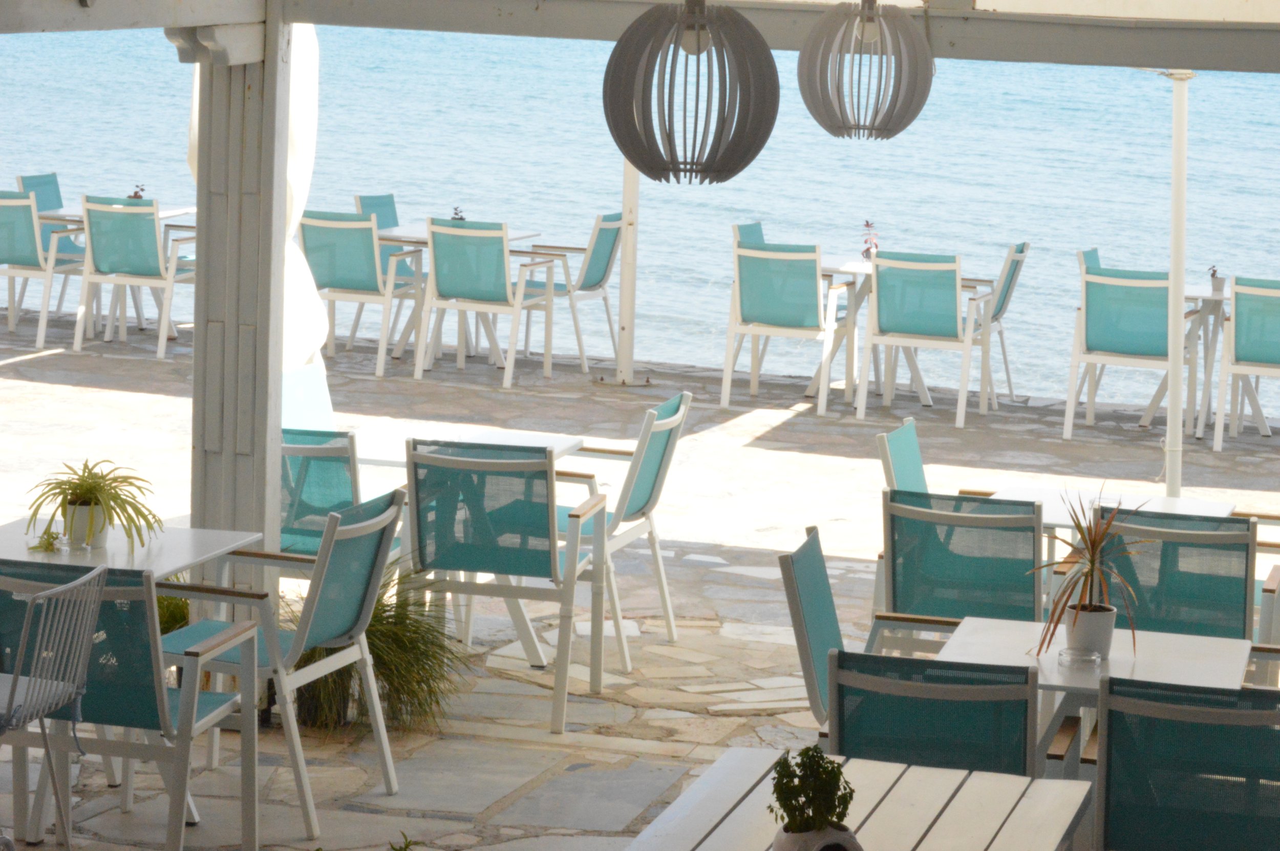 Gelateria Maria - Outdoor seaside restaurant with white tables and turquoise chairs, overlooking the calm water, decorated with potted plants and hanging lantern lights.