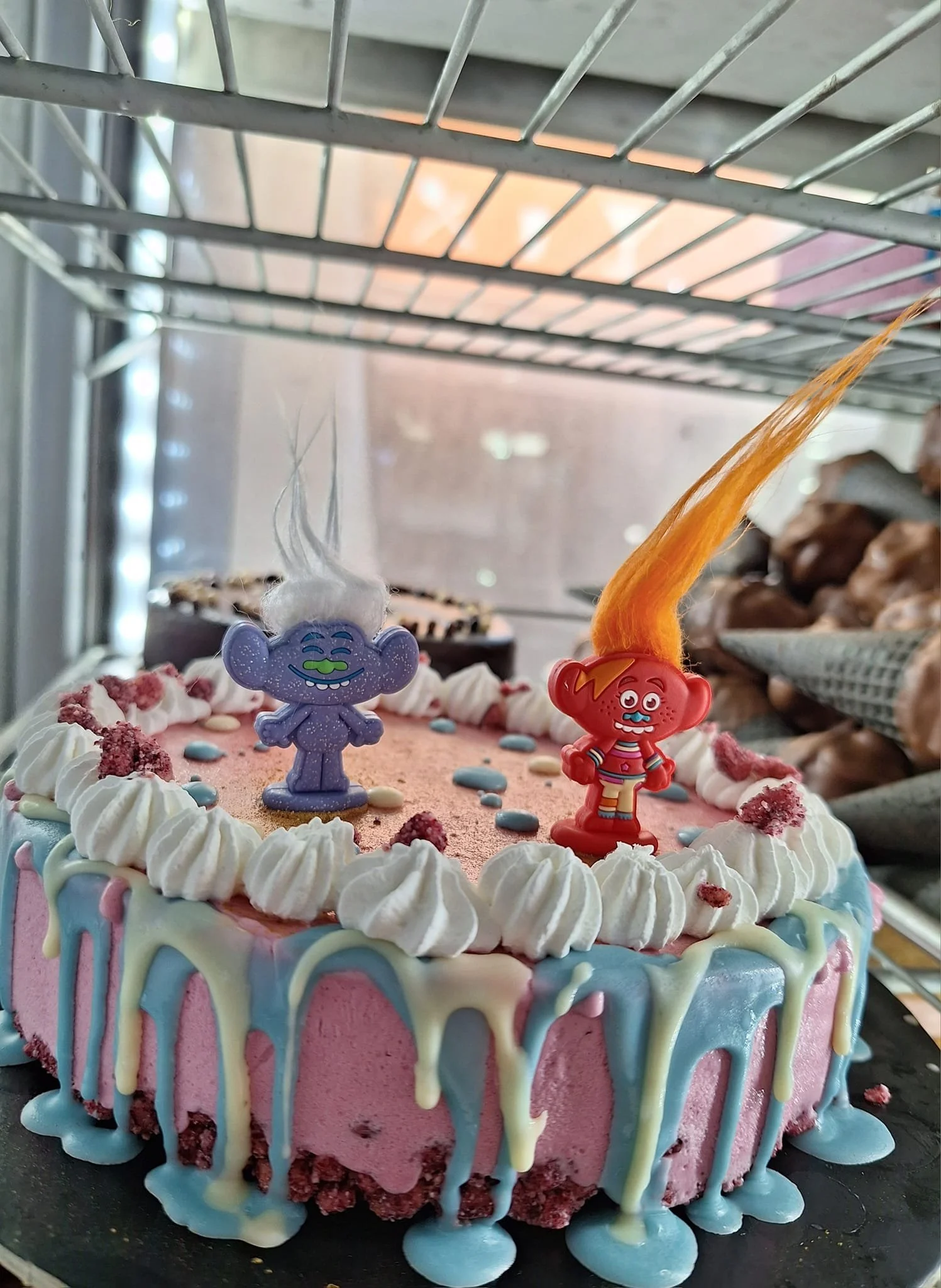 Gelateria Maria - Colorful birthday cake with pink, white, and pastel blue icing, decorated with doll toppers with hair made of cotton candy, in a bakery display case.