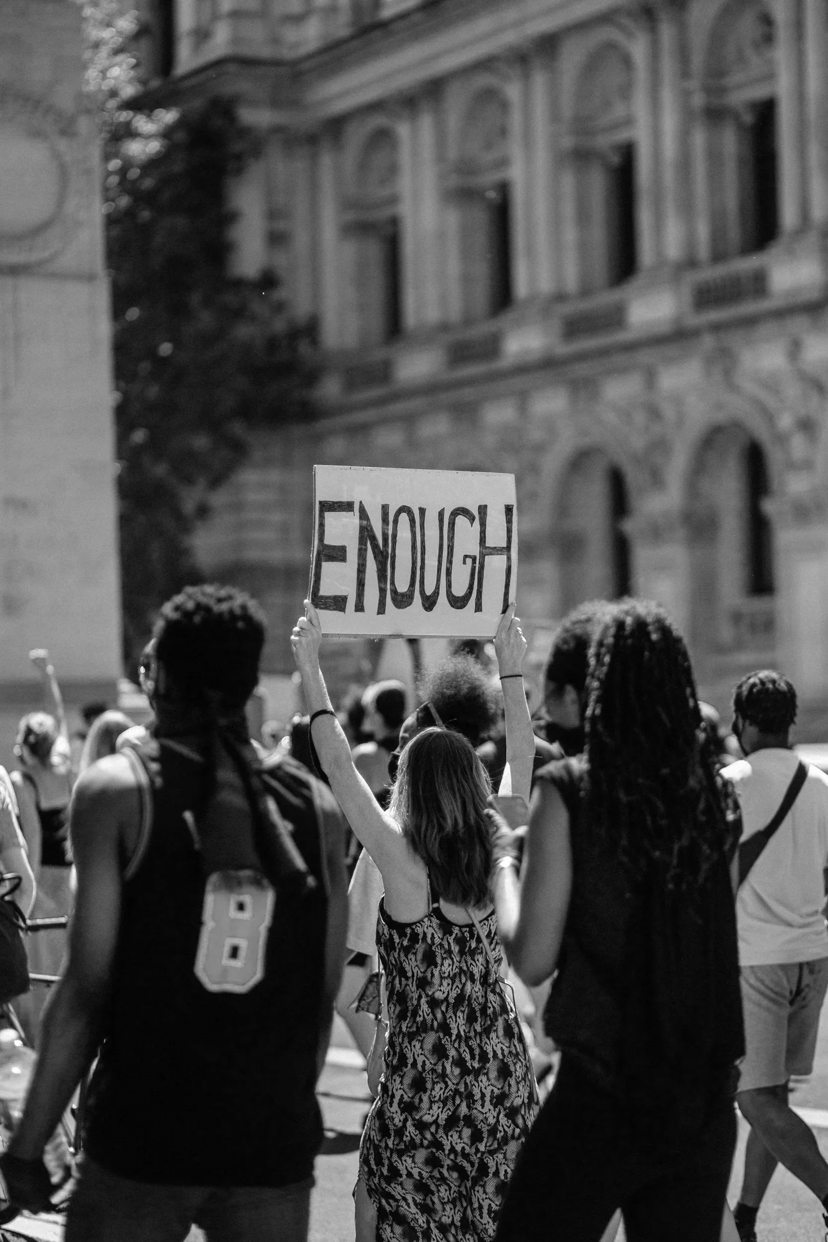 People participating in a protest march, with one person holding a sign that reads "ENOUGH." The scene takes place outdoors in front of a historic building, possibly a government or courthouse building, during the daytime.