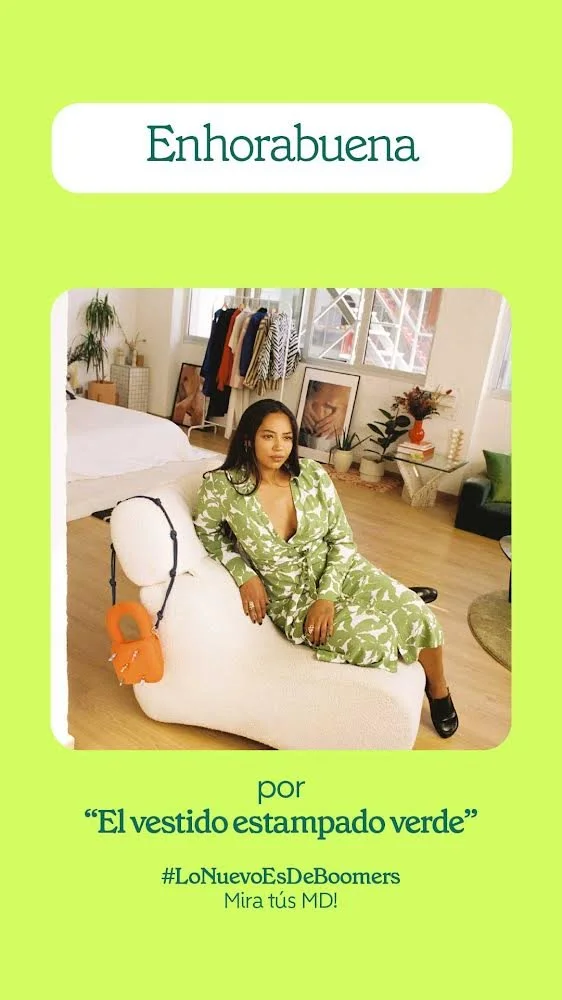 A woman wearing a green printed dress sitting on a white plush chair in a bright, modern room with large windows, plants, artwork, and clothing racks in the background.