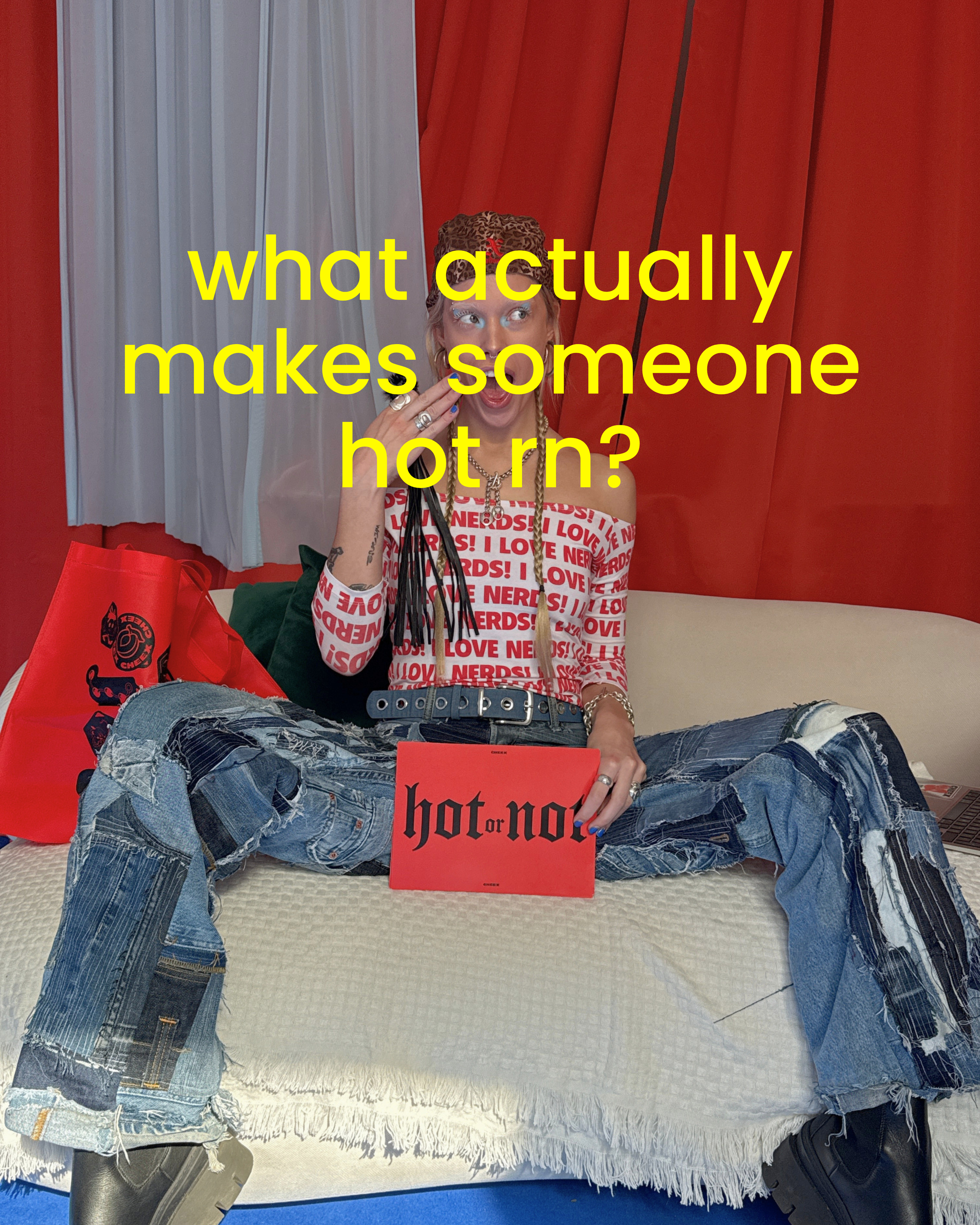 A person sitting on a bed with legs crossed, wearing ripped jeans and boots, holding a red book that reads 'hot: or not'. They are dressed in a long-sleeve top with red and white patterns that say 'LOVE NERDS!', and accessorized with rings, a necklace, and a hat with leopard print. There are red shopping bags on the bed, and behind them are red curtains with some white curtains on the side.