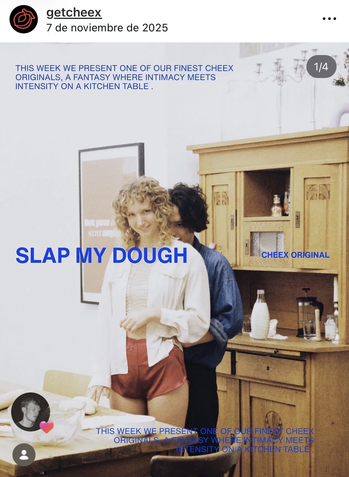 A woman with curly blonde hair and a man with black curly hair stand in a kitchen, smiling. The woman is dressed in a white top and red shorts, while the man is in a denim shirt. The kitchen features a wooden cabinet, bottles, and a kettle. The image